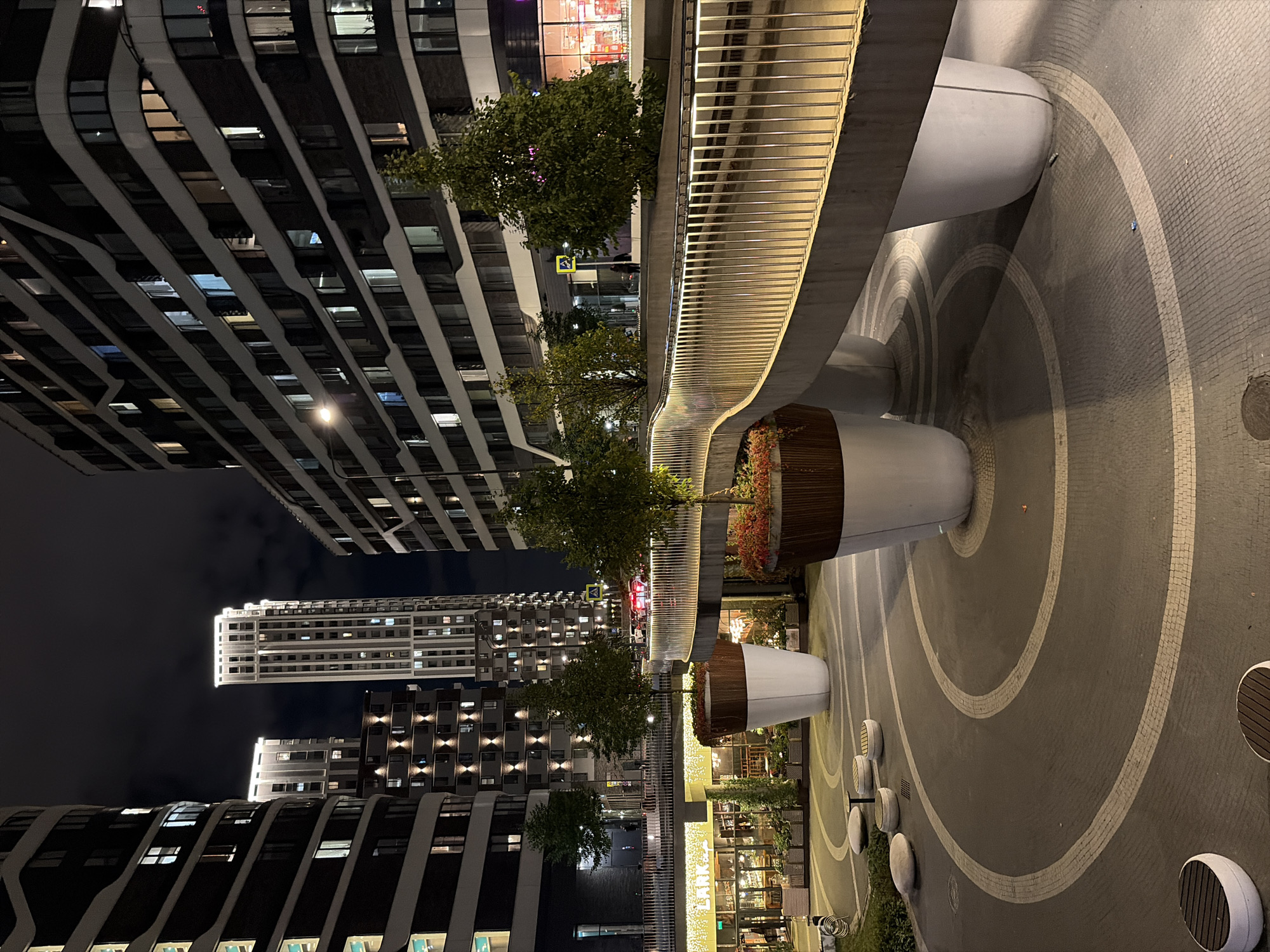 The image shows an outdoor urban scene at night, featuring modern architecture and a pedestrian walkway. The buildings are tall and contemporary, with many illuminated windows, indicating residential or office spaces. The walkway itself is elevated and curves through the scene, with a sleek metal railing illuminated by embedded lights. Large, white planters containing trees and plants are strategically placed around the walkway and in the foreground. The ground in the foreground has a circular pattern of light and dark paving stones, with some rounded benches and planters. In the background, there are glimpses of businesses with illuminated storefronts. The sky is dark and cloudy. The overall impression is of a modern, well-lit public space in a city. This scene is located in Moscow, Russia, specifically in the residential complex "Domodedovo" on the Kashirskoye Highway, a district known for its modern high-rise buildings and redeveloped public spaces.