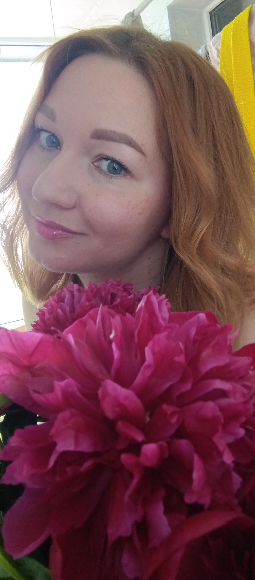 A woman with fair skin and reddish-brown hair smiles at the camera, holding a bouquet of large, bright pink flowers that partially obscures her body. She has light blue eyes and is wearing pink lip gloss. The background includes a light-colored wall and a hint of a yellow garment or object. The location is unknown.