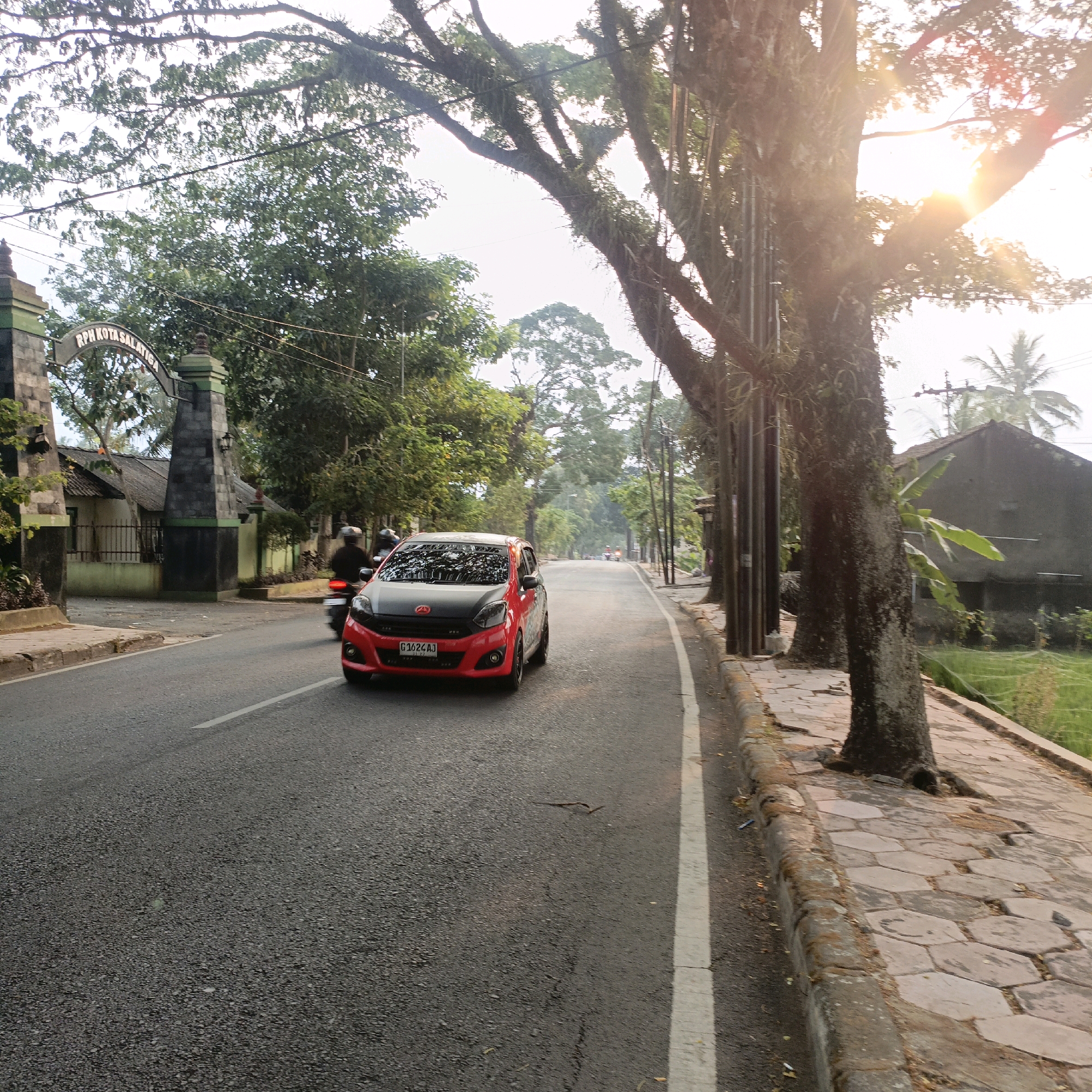 The image depicts a street scene in Salatiga, Indonesia, likely during the late afternoon or early morning, given the warm sunlight and lens flare. In the foreground, a red and black car with license plate G 1624 AJ is traveling on an asphalt road. To the left of the car, a motorcycle with two riders is also moving in the same direction. In the background, a large, old tree with thick branches laden with epiphytes dominates the right side of the frame. The street is lined with a paved sidewalk and a grassy area. Further back, an arched gate with the inscription "RPH KOTASALATIGA" is visible, along with a building and more trees. The overall impression is that of a semi-urban or suburban environment with significant natural elements.