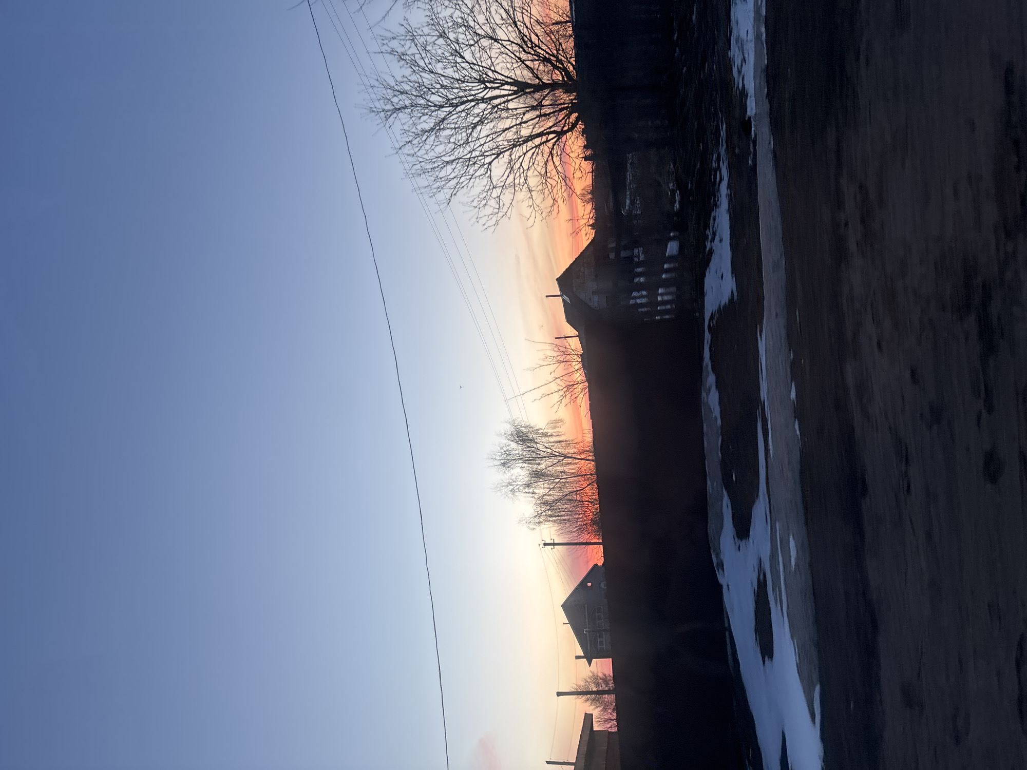 The image depicts a twilight or dawn scene in what appears to be a rural or semi-rural area of Ольховка, Russia. The sky transitions from blue to orange/red, indicating the presence of the sun near the horizon. Several buildings are visible, silhouetted against the colorful sky. Bare trees line the horizon. Utility poles and wires are visible. A partially snow-covered area is in the right foreground. The scene lacks any clear indication of human presence or activity.