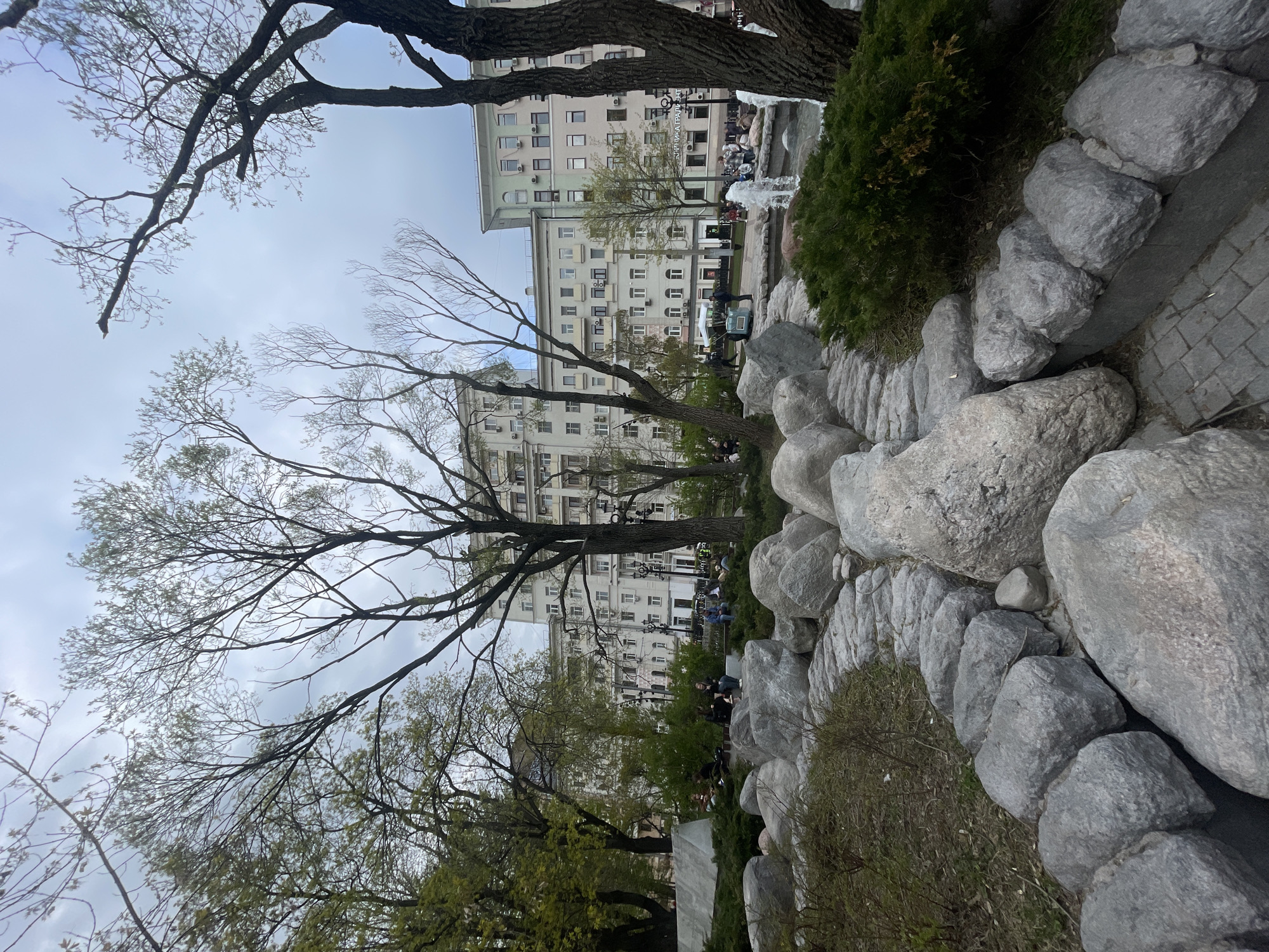 The image depicts a park scene in Moscow, Russia. The foreground features a rock formation bordering a paved walkway. Trees with budding leaves are interspersed between the rocks. Behind the trees is a multi-story building. People are situated near a fountain in the background. The sky is overcast.