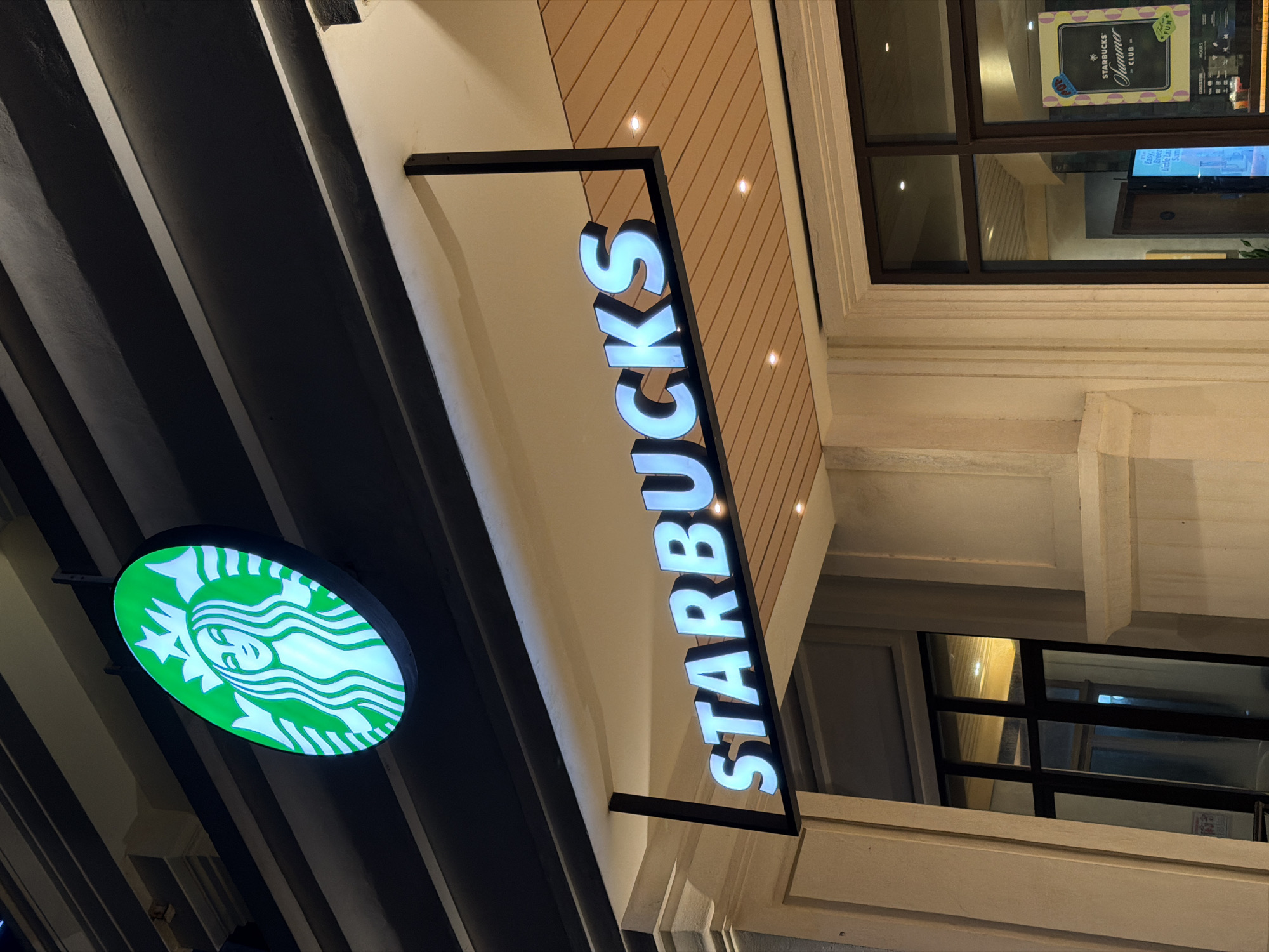 This media file is a nighttime photograph of a Starbucks storefront in Da Nang, Vietnam. The main subjects are the illuminated Starbucks logo, a circular sign with the iconic siren in green and white, and the vertically oriented "STARBUCKS" lettering in bright white. The storefront features a facade with wooden-paneled accents above the windows, and cream-colored architectural elements. Dark, angled beams are visible to the left of the signage. Inside the store, visible through the windows, are reflections of lights and what appears to be a promotional poster for Starbucks' "Summer" collection, featuring the text "STARBUCKS Summer by CLUB" and what looks like a flower graphic. The overall impression is that of a modern, well-lit commercial establishment operating in the evening. The time of day is clearly night due to the artificial lighting and the dark exterior. There are no people or specific weather conditions visible. The location is identifiable as Da Nang, Vietnam.