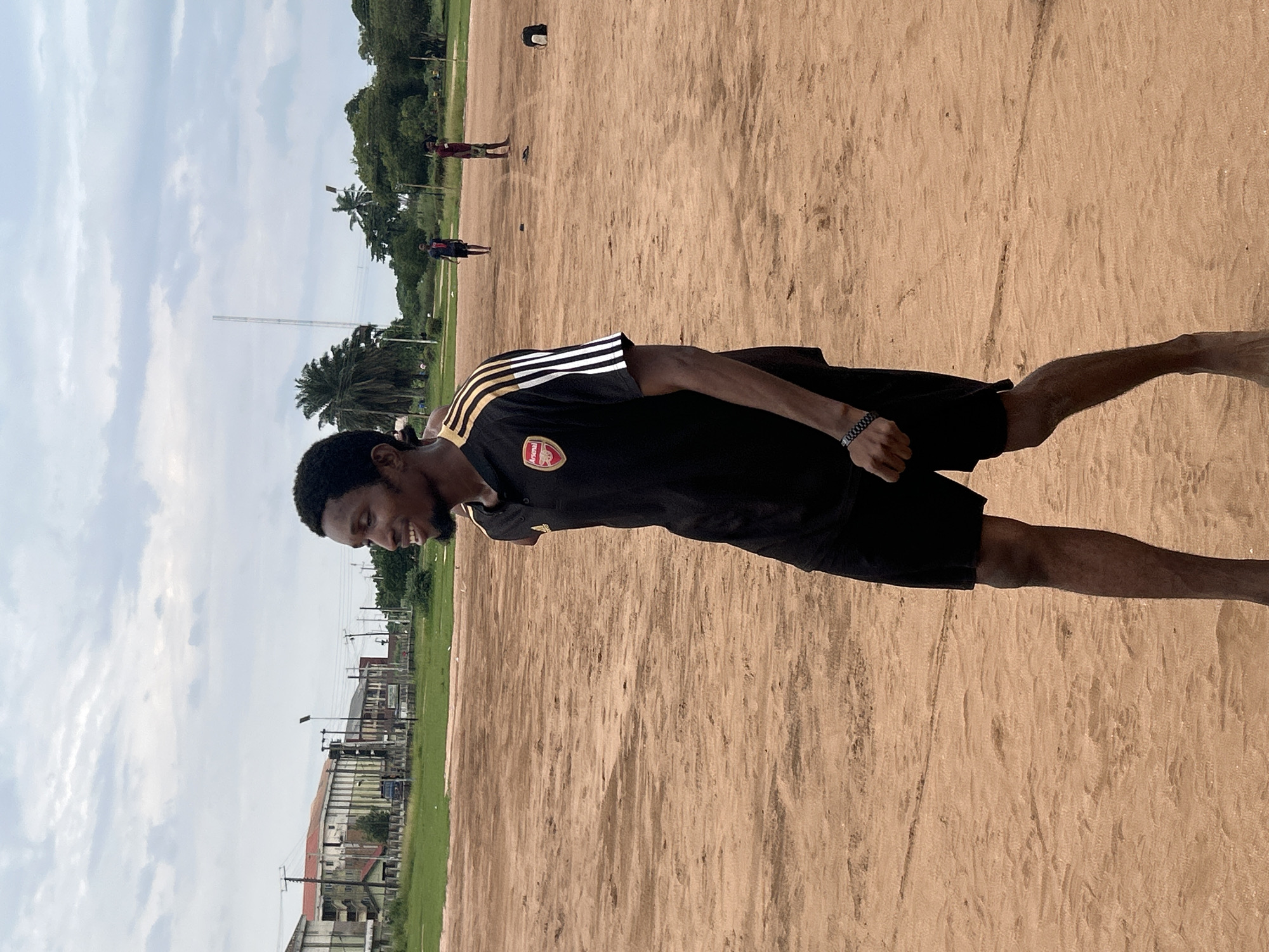 In Ihiagwa, Nigeria, a young Black man stands on a sandy field. He is wearing a black t-shirt with gold stripes on the sleeves and the Arsenal logo on the chest, along with black shorts and a black bracelet. He is barefoot and appears to be smiling and looking to his left. The field is sandy with some patches of dry grass. In the background, there are trees, buildings, and a cloudy sky. Two other figures are visible in the distance on the field, and a black backpack lies on the sand.
