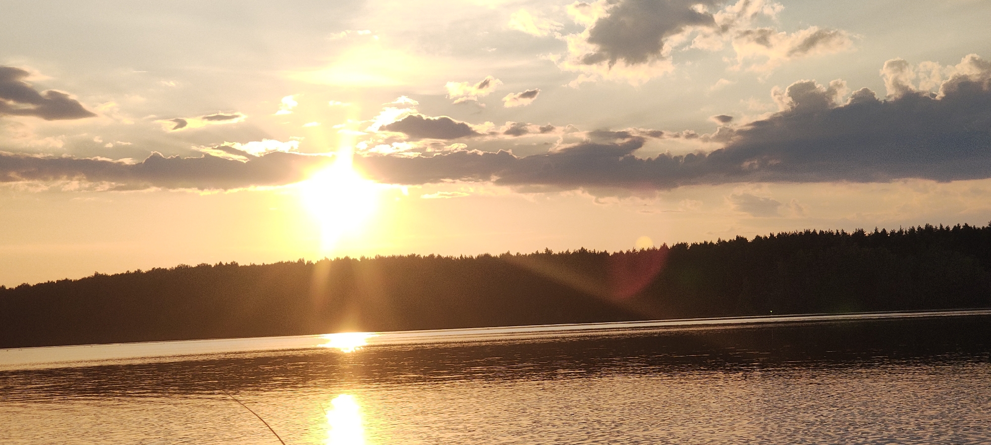 The image depicts a sunset over a body of water, likely a lake, with a treeline forming a dark silhouette against the vibrant sky. The sun is a bright, central focus, casting golden rays across the scene and reflecting brilliantly on the water's surface. The sky is a mix of warm orange and yellow hues, with scattered clouds, some darker and more defined, others lighter and wispy, tinged with the sunset colors. The water is calm, with gentle ripples visible, also reflecting the dramatic sky. The treeline appears dense and suggests a natural, undeveloped landscape bordering the water. The overall impression is one of natural beauty and tranquility at dusk. The location in Mednoye Ozero, Russia, implies a serene natural setting commonly found in such regions during this time of day.