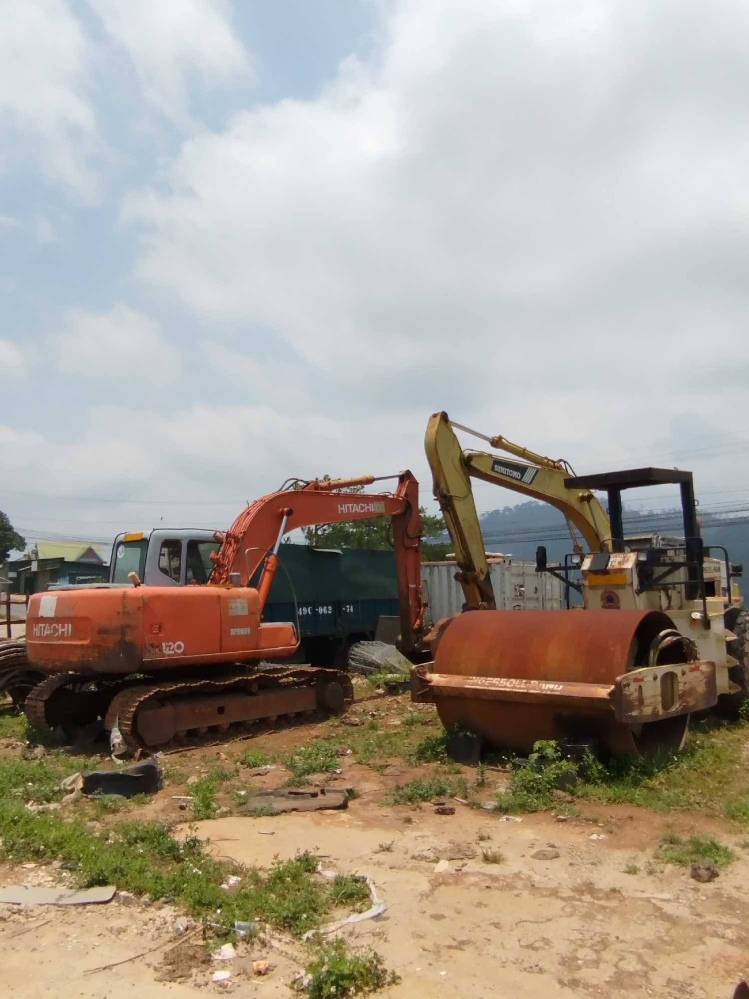 The image shows two pieces of heavy machinery in a lot. An orange Hitachi excavator with a visible "120" model number is on the left. To the right of the excavator, there is a yellow Sumitomo road roller with a rusted drum. The ground is dirt with patches of grass. In the background, there appears to be a green truck, some white storage containers, and some indistinct buildings and foliage. The sky is partly cloudy. The location is estimated to be in Păng Tiêng 1, Vietnam, based on the prompt. There are no people visible in the image.