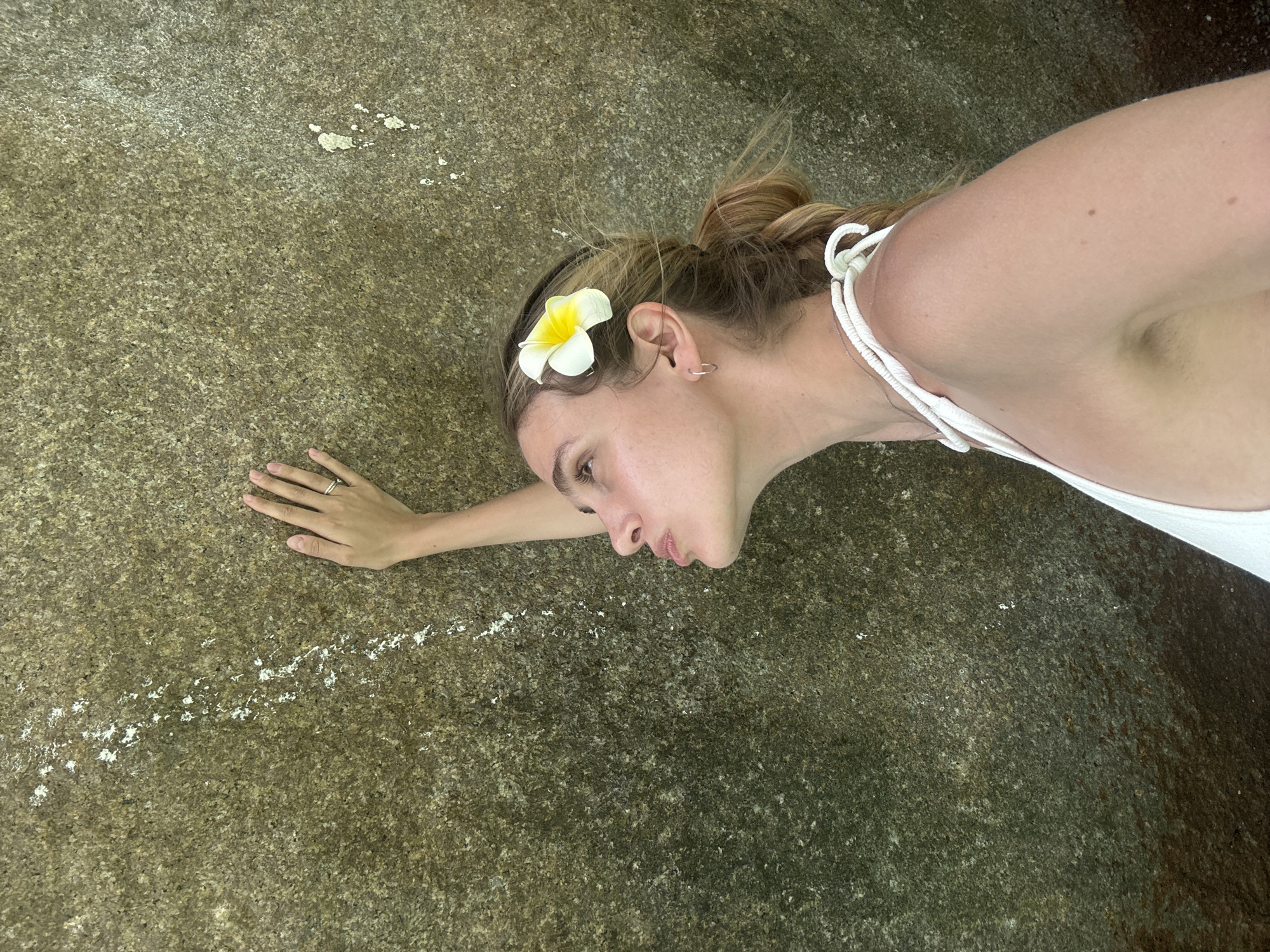 A fair-skinned woman lies on a rough, textured rock surface. A white and yellow flower is positioned in her hair above her left ear. She wears a white top with thin straps. Her left hand rests on the rock, displaying a ring on her ring finger. The location is likely near Lam Kaen, Thailand, given the tropical flora. The rock has a textured, mottled pattern with some white deposits along a linear path.