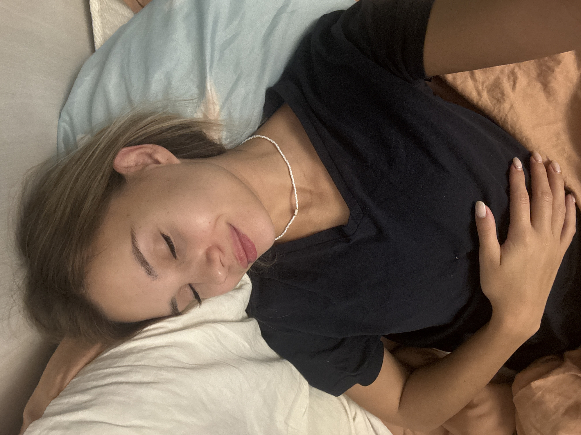 The image depicts a young woman resting comfortably in what appears to be a bed, likely indoors. She is lying on her side, facing slightly away from the camera, with her head on a white pillow and her hair spread around her. Her eyes are closed, and a gentle, peaceful smile is visible on her lips, suggesting a state of relaxation or sleep.
The woman has light brown or blonde hair and fair skin. She is wearing a simple black, short-sleeved t-shirt. A delicate, thin white beaded necklace with a few rectangular beads adorns her neck. Her left hand rests lightly on her torso or the bedding, revealing nails painted with a light, neutral polish.
The bedding includes a light blue pillow or blanket behind her head and a white sheet or pillow beneath her. A peach or tan-colored blanket is visible in the background, contributing to the soft, warm tones of the setting.
The scene conveys a calm and intimate atmosphere, possibly during nighttime or an afternoon nap. There are no discernible location cues within the image itself to confirm it is Chaikovsky, Russia, nor is any text visible. The overall mood is serene and tranquil.