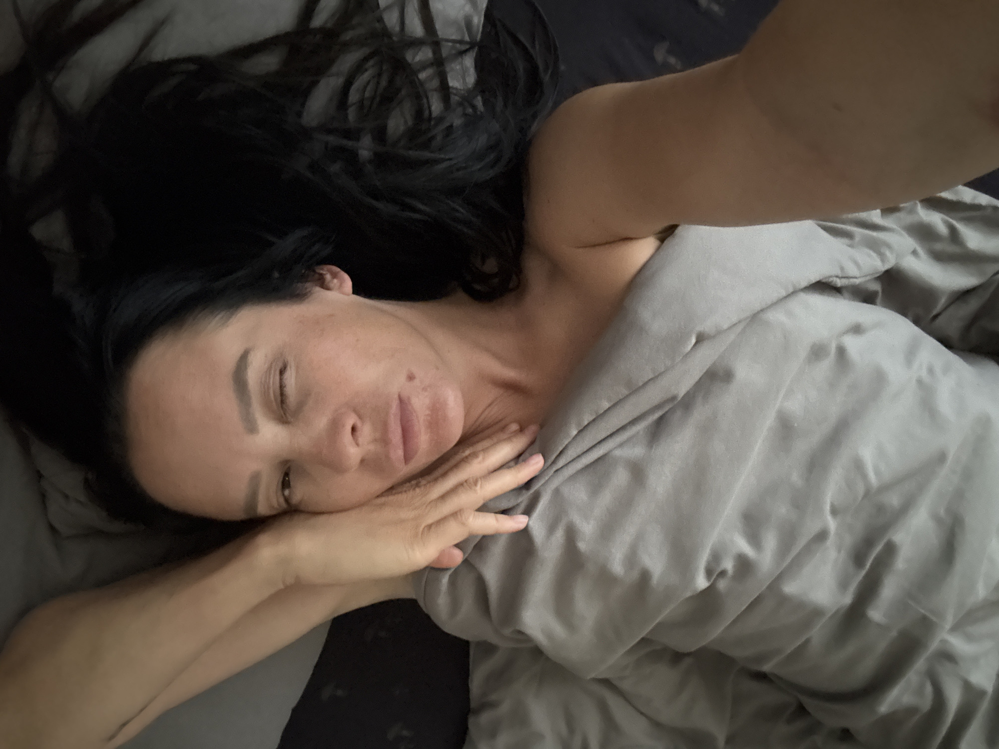 A woman with dark hair is lying in bed under a gray blanket. She is partially covered by the blanket, and her right arm is stretched out towards the camera. Her eyes are partially closed. The scene is dimly lit. There is no evidence to suggest that this image was taken in Chaikovsky, Russia.