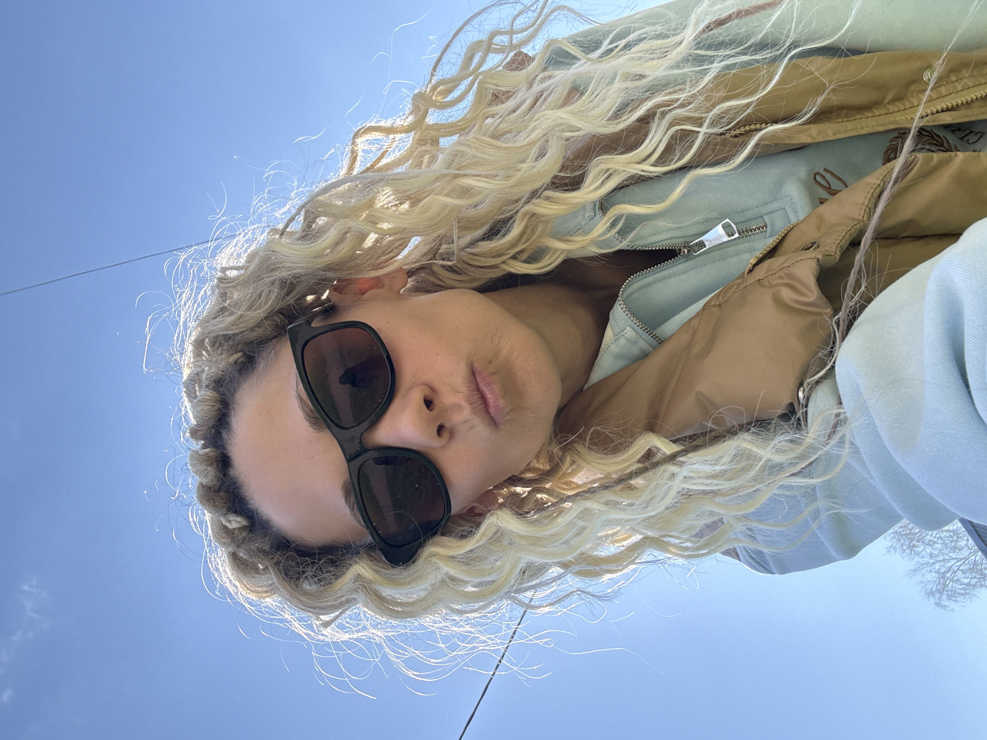 A low-angle, upward-facing photograph depicts a person with light blonde, wavy hair, wearing black sunglasses. The person's head is tilted back, and the clear blue sky is visible in the background. The person is wearing a light blue jacket with a tan lining, with a zipper visible. Thin, dark lines, possibly power lines, cut across the upper portion of the frame. Bare tree branches are visible in the upper right corner. The location is presumed to be Rusanovo, Russia.