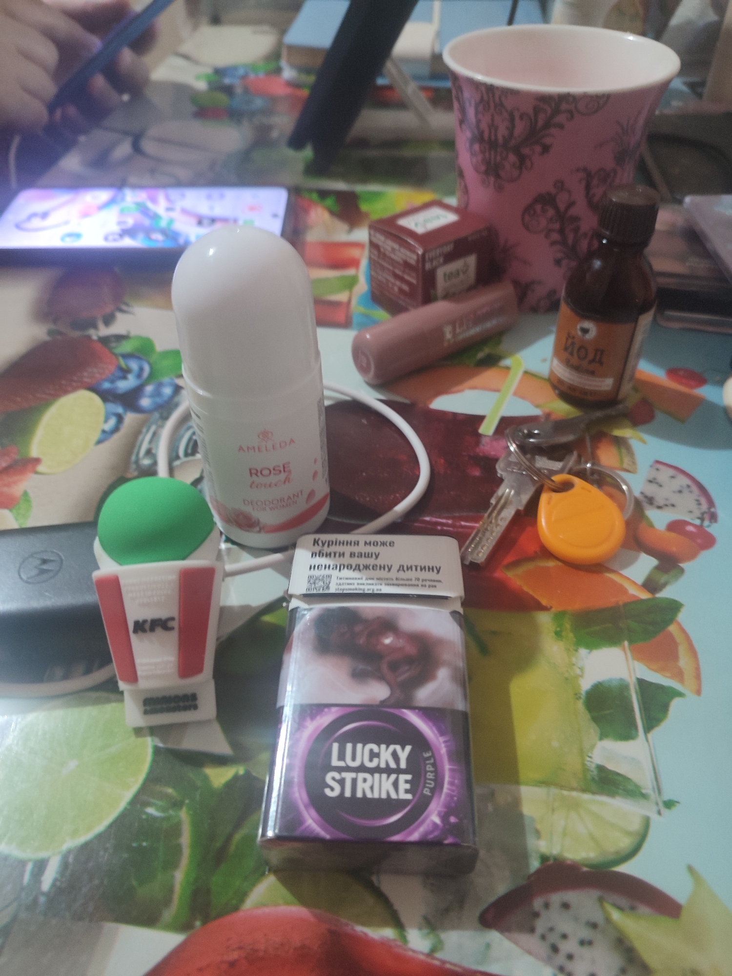 The image depicts a table covered with a colorful cloth adorned with fruit and beverage designs, likely in a casual setting. Several objects are scattered across the surface. In the foreground, a pack of "Lucky Strike" cigarettes, specifically the "Purple" variant, is prominent. To its left, a white roll-on deodorant with the brand "AMELDA" and the scent "ROSE touch" is visible. Next to the deodorant is a miniature red and white KFC bucket, possibly a novelty item or holder, with a green sphere atop it.
Further into the scene, a small brown glass bottle containing a liquid labeled "ЙОД" (iodine) stands near a pink decorative cup with a black damask pattern. A lipstick in a muted pink shade lies horizontally. A red cardboard box, possibly containing tea, is also present. In the background, a smartphone with a colorful display is partially visible, and a person's hands are manipulating a dark-colored object, possibly another phone or tablet. A set of keys with an orange fob is also on the table, along with a portion of a laptop. The overall impression is one of everyday items casually placed on a table, suggesting a personal space or a moment of relaxation. The specific location within Chernivtsi, Ukraine, cannot be determined from the visual information alone.