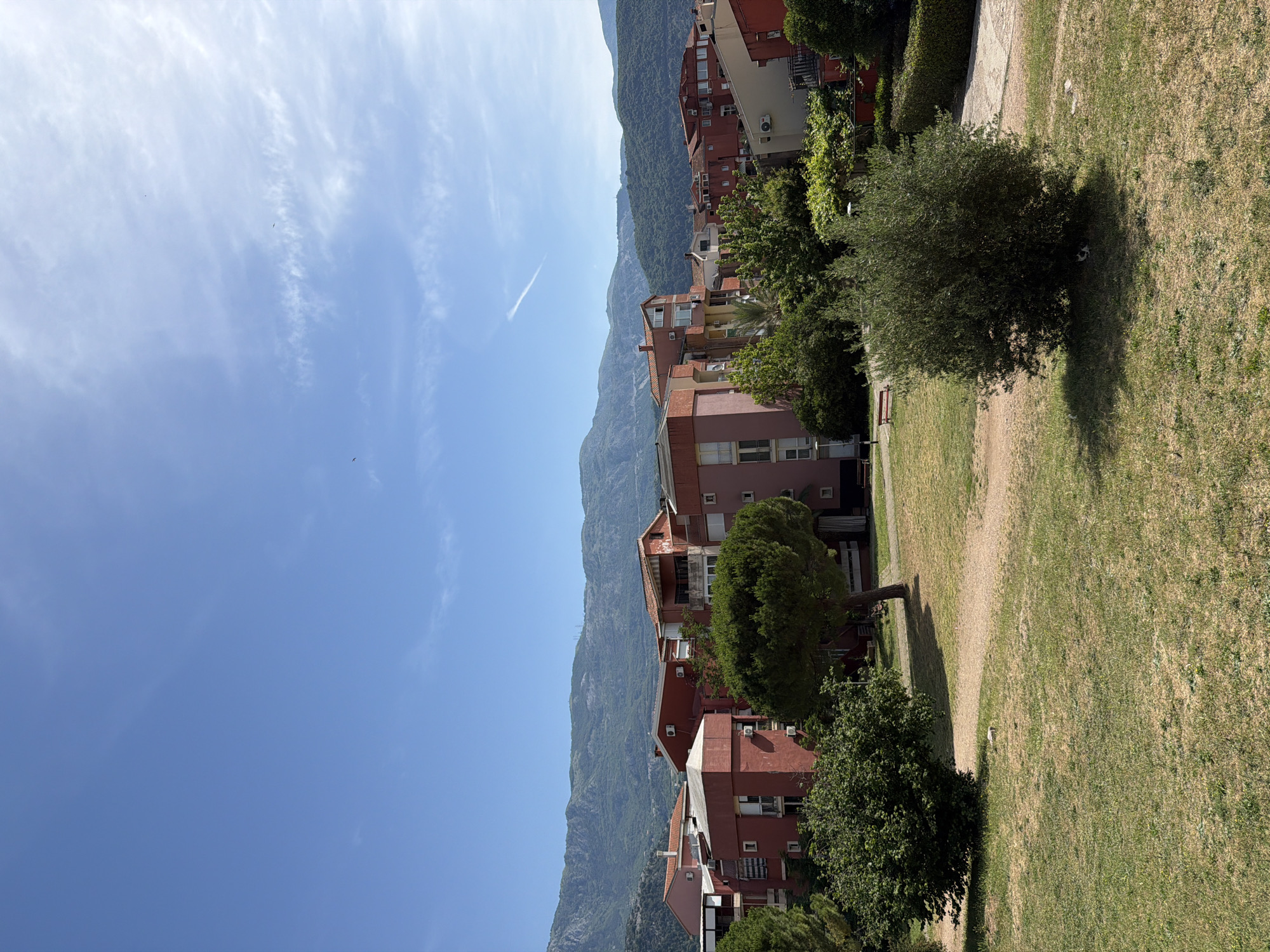 The image depicts a residential area in Budva, Montenegro. The scene includes red-roofed buildings nestled near mountainous terrain. A green lawn with scattered trees and a pedestrian pathway occupy the foreground. The sky is a clear blue with a few wisps of clouds and what appears to be a contrail from an airplane. No people are visible.