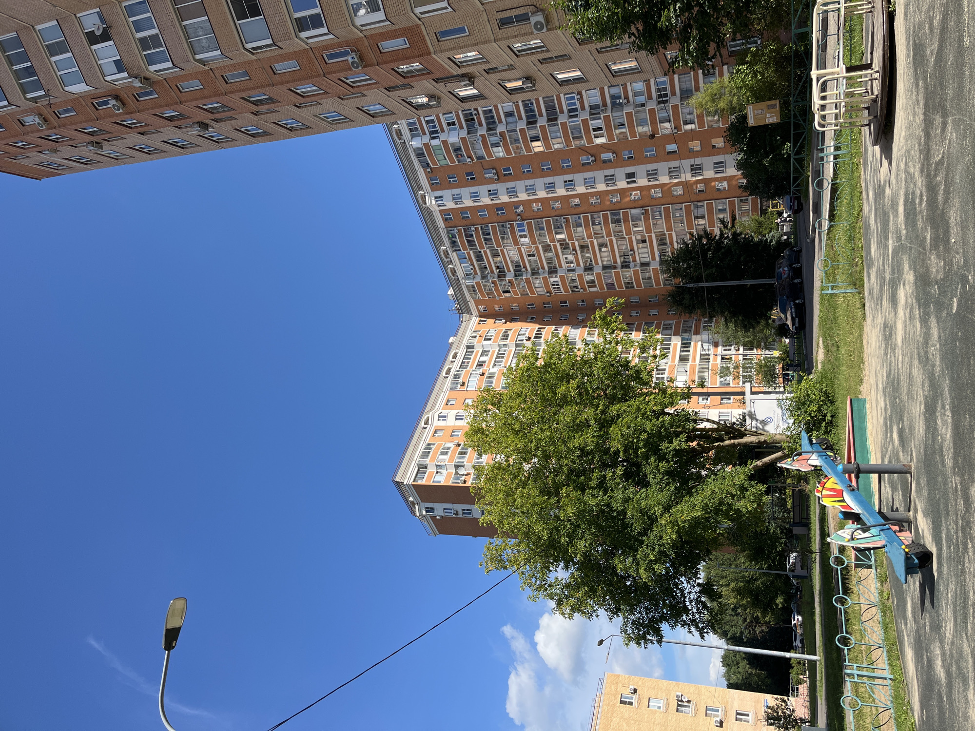The image captures an outdoor scene in the city of Balashikha, Russia, on a clear, sunny day. The foreground features a playground with a blue seesaw and a merry-go-round. A light blue metal fence with circular accents surrounds a grassy area. In the middle ground, a large, leafy green tree stands prominently, with more trees and bushes visible behind it. Several cars are parked along a street that runs behind the playground. The background is dominated by the presence of tall, multi-story apartment buildings, characterized by their orange and light-colored brick facades, numerous windows, and balconies. One building is particularly large and curves around a portion of the scene. A street lamp is visible on the left side of the image, and power lines stretch across the clear blue sky. The environment appears to be a residential urban area.