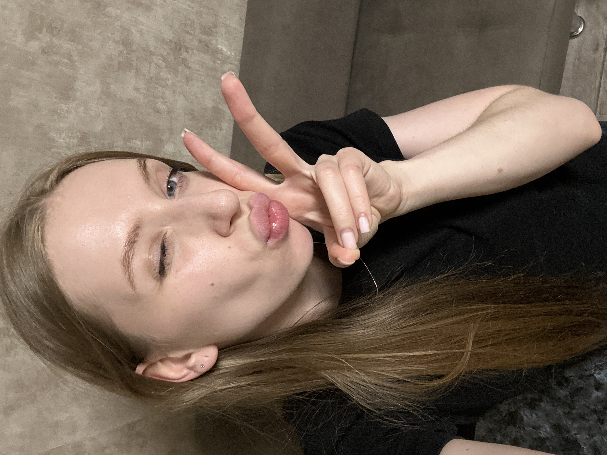An indoor, close-up, selfie-style portrait captured in what is stated to be the city of Chaikovsky, Russia, features a young adult female from the upper torso upwards. The subject has long, straight, light brown hair, parted on the left, partially obscuring the left shoulder. Her left eye is open and blue, exhibiting eyeliner, while her right eye is closed in a wink. Her lips are pursed into a "kissing" gesture, appearing glossy. The left ear is visible, displaying two small, silver-toned stud earrings in the lobe. Her left hand is raised, performing a "peace sign" gesture with the index and middle fingers extended; the fingernails are manicured with light pink polish. She is wearing a black short-sleeved t-shirt, and a thin, gold-colored necklace chain is partially visible around her neck. The background consists of a textured, light beige wall on the left and a portion of a dark brown or grey upholstered sofa on the right. The lighting suggests an indoor environment. No specific city identifiers are visible within the image itself.