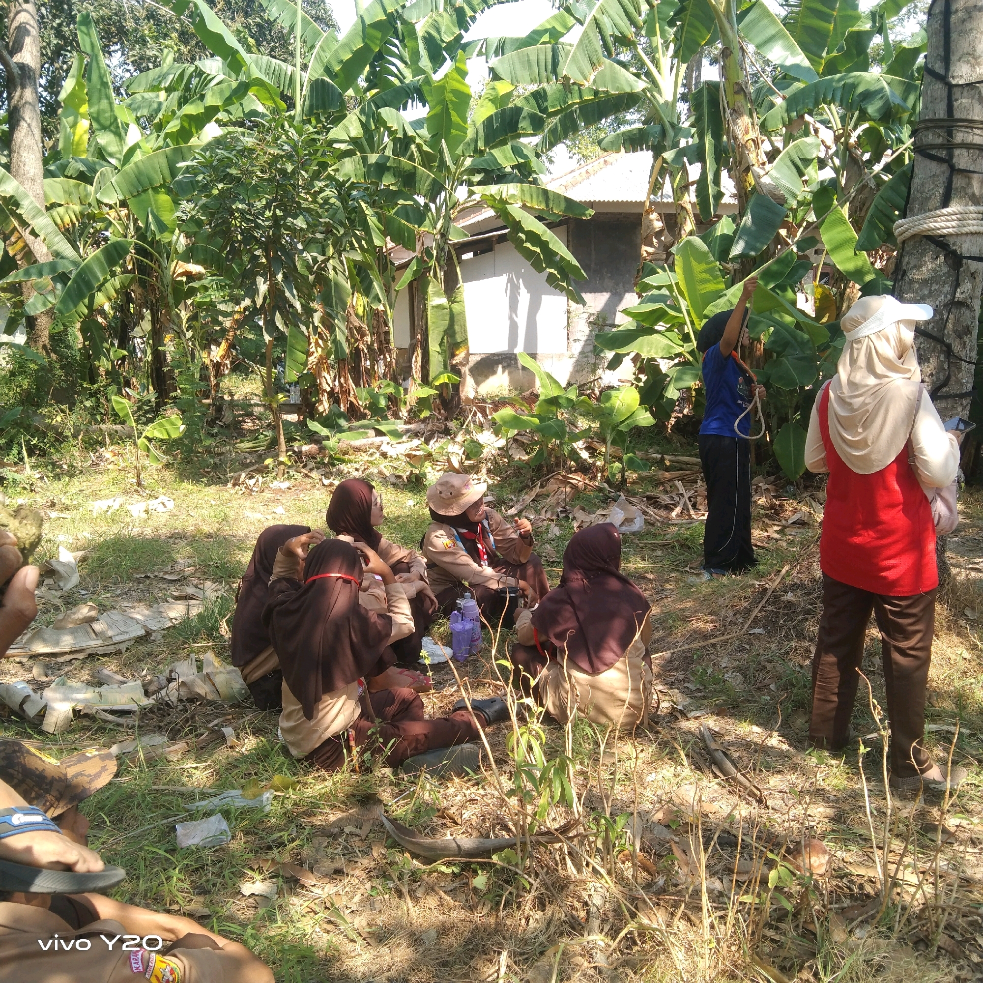 In Karawang, Indonesia, a group of individuals are gathered outdoors amidst lush banana trees and vegetation. Several people, dressed in brown scout uniforms and hijabs, are seated on the ground. One individual in the foreground is wearing a light brown hijab and is looking to their left. Another person in a scout uniform with a hat is seated and appears to be speaking. To the right, two individuals are standing, one wearing a red vest over a light-colored hijab and the other in a blue shirt and black pants, holding a rope. The environment is natural, with sunlight filtering through the leaves, casting shadows on the grassy, somewhat unkempt ground. There are scattered dry leaves, twigs, and some plastic water bottles on the ground. The scene suggests an outdoor activity or gathering, possibly related to scouting.