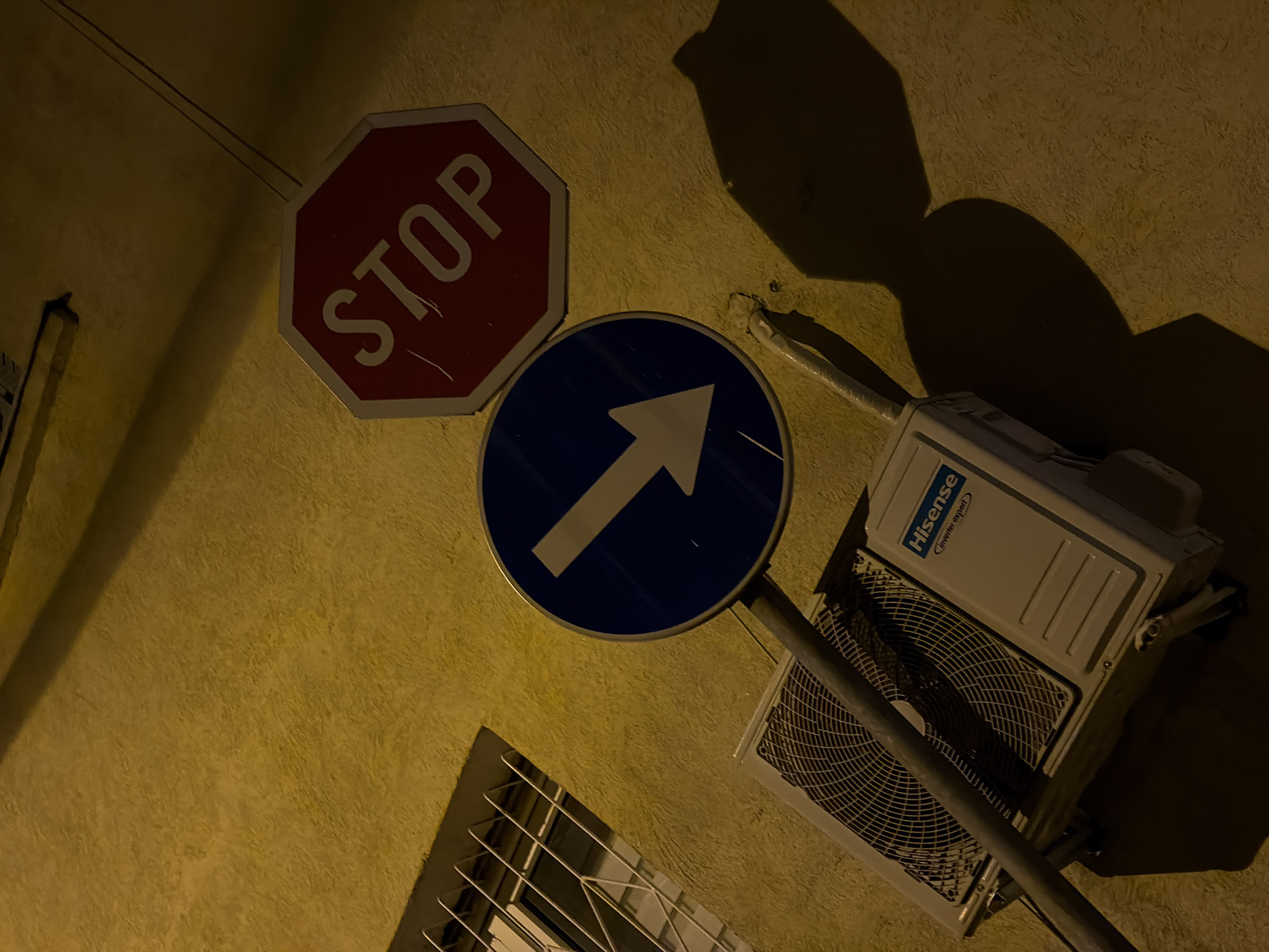 The image depicts a section of a building wall in what is likely Belgrade, Serbia, due to the presence of Cyrillic script and architectural styles common in the region. Attached to the wall are two traffic signs: a red octagonal "STOP" sign and a blue circular sign with a white arrow pointing upwards, indicating a one-way street. Below the signs is a window with a metal security grill. To the right of the signs is a Hisense brand air conditioning unit mounted on the wall. The wall appears to be textured and is illuminated by a warm light source, casting shadows of objects on the wall. No people are visible. The scene is stationary and devoid of any clear action or interaction.
