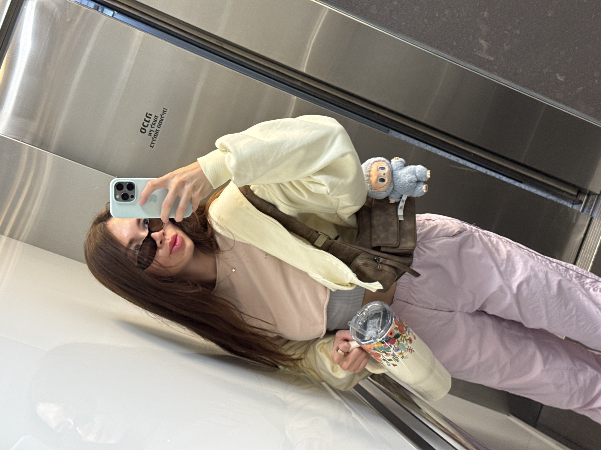 A young woman is taking a selfie in what appears to be an elevator. She is wearing sunglasses, a cropped light pink top, a light yellow zip-up hoodie, light purple pants, and a brown crossbody bag with a small stuffed animal attached. She is holding a smartphone in her left hand and a floral-patterned tumbler in her right hand. The elevator's interior is stainless steel. A sticker with the Hebrew letters "OCLG" is visible in the upper left area of the elevator. Based on the "OCLG" sticker, the elevator is in Ramat Gan, Israel.