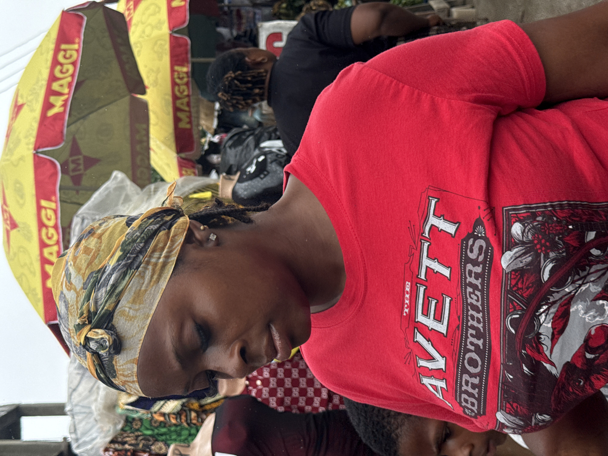 In Lekki, Nigeria, a woman wears a red t-shirt with "The Avett Brothers" printed on it and a yellow and green headwrap. She stands in front of stalls with yellow Maggi umbrellas. Another person, partially visible on the left, wears a dark garment. In the background, a person with braided hair wears a black t-shirt. The environment appears to be an outdoor market on a cloudy day.