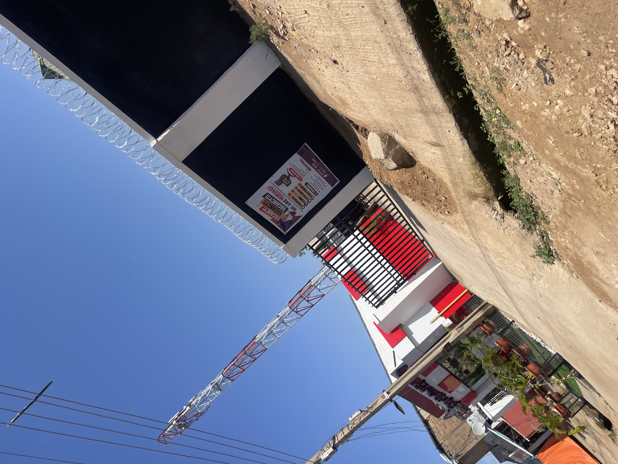 The image shows a construction site in Gura Top, Nigeria, under a clear blue sky. In the foreground, a concrete ditch runs diagonally across the frame, with dirt and rocks piled on its edges. To the left, a utility pole with wires stretches upwards. Behind the pole, a red and white building with modern architecture is visible, featuring red accents and a black and red gate. Further to the left, a large orange tarp is draped over an unseen object. In the middle ground, a red and white lattice tower stands tall, reaching towards the sky. Behind the building and to the right, a black wall is topped with barbed wire fencing. A banner on the wall advertises "VACANCY! WE ARE HIRING!" listing various positions and contact information. Potted plants are arranged in front of the building. The ground is uneven, with patches of dirt and concrete.
