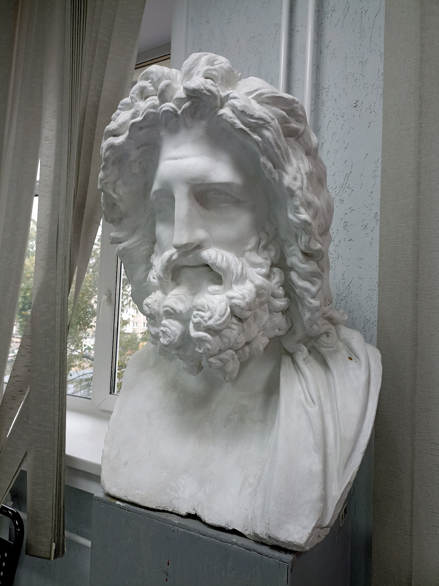 A white plaster bust of a bearded man with curly hair is positioned in front of a window. The bust is placed on a grey pedestal. To the left of the bust, a light-colored curtain is drawn, partially obscuring the window. Outside the window, trees with autumn foliage and buildings are visible. The wall behind the bust is textured and light-colored, with vertical decorative elements. A grey fabric is on the right side of the image. The overall scene appears to be indoors, in an academic or art studio setting, given the nature of the bust.