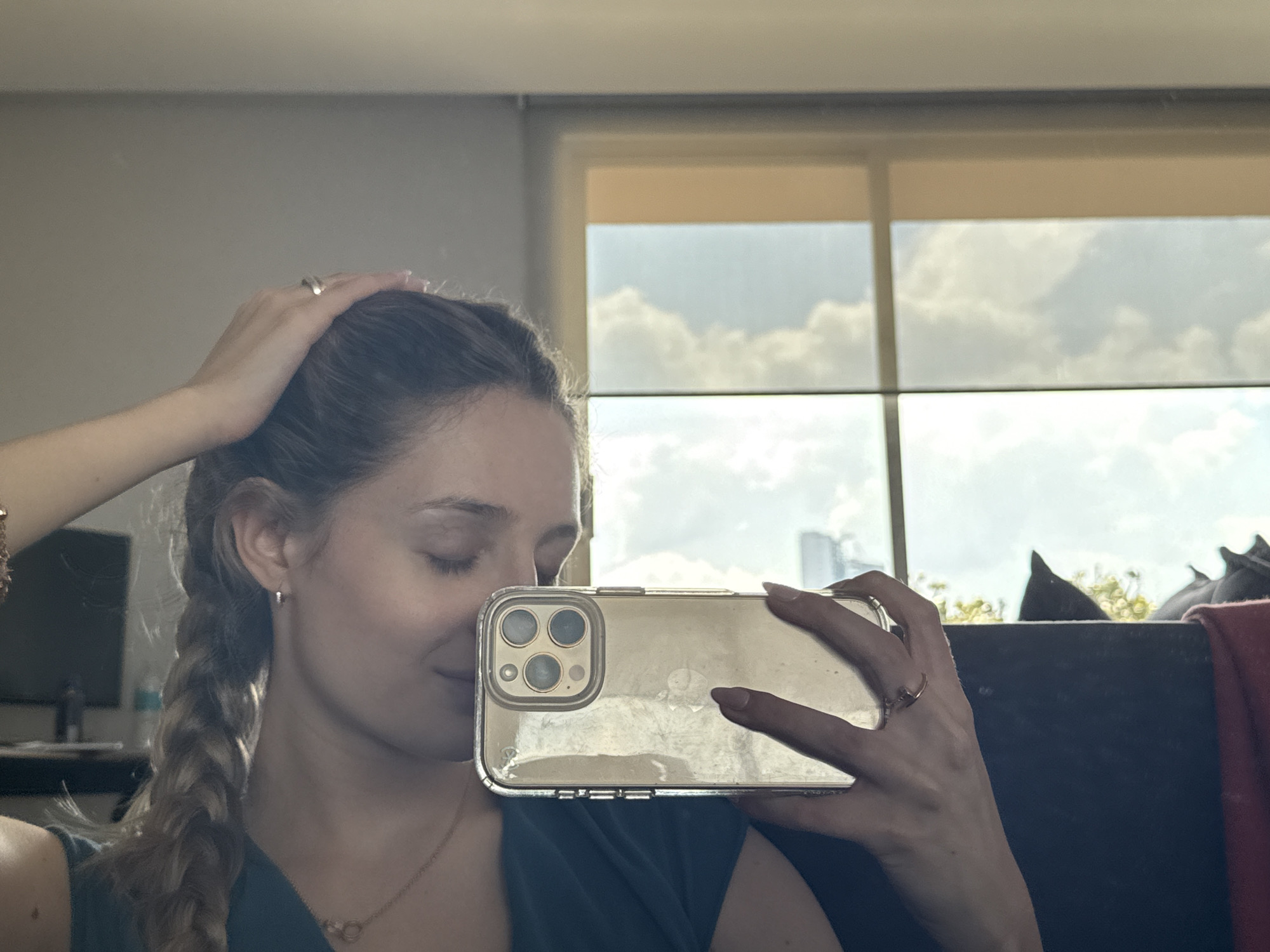 A young woman is captured in a mirror selfie, showing her from the chest up within an indoor setting. She has long, light brown hair, styled partly in a braid over her left shoulder, and is wearing a dark-colored top. Her left hand is raised to her head, touching her hair, and a ring is visible on her right ring finger, which is holding a smartphone. The phone is encased in a clear cover, showcasing a triple-lens camera system, and reflects bright light.
The woman's eyes are closed or cast downwards, and a gentle smile plays on her lips, suggesting a relaxed or content mood. She wears a necklace featuring two interlocking circular pendants and small hoop earrings.
In the background, visible through a large window, is a bright, cloudy daytime sky. Distant buildings or a city skyline are faintly discernible, consistent with an urban environment like Mexico City. The window also has a light-colored blind or shade pulled down slightly from the top. On the far left of the mirror's reflection, a television or monitor is visible on what appears to be a desk or shelf. No visible text is present in the image.