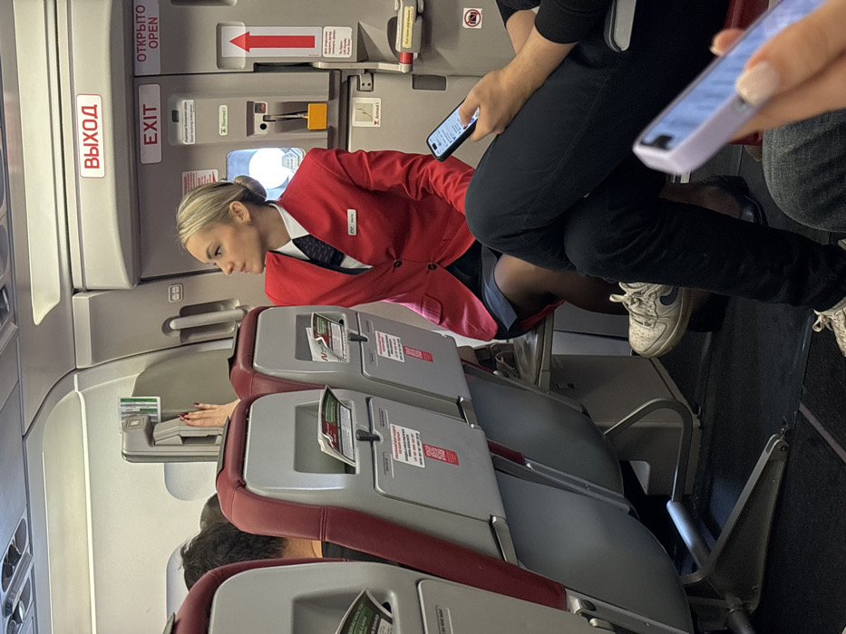 The image displays the interior of an aircraft cabin, located in the city of Novogorelovo, Russia.
A blonde female flight attendant, dressed in a red blazer, white collared shirt, and dark tie, is seated in the mid-ground, facing left and slightly upwards. Her right hand is extended towards an overhead panel or mechanism located above the emergency exit door. The exit door, positioned to her right, features both Cyrillic and English labels, including "ВЫХОД" and "EXIT," along with a red arrow.
In the foreground, the lower body of a passenger is visible, wearing dark trousers and white sneakers, with their legs crossed. This individual is holding a smartphone in their right hand. Adjacent to this, partially visible on the far right, is another passenger's hand holding a smartphone with a light purple case.
The cabin interior features rows of aircraft seats upholstered in red and grey, equipped with seatback pockets. The ceiling displays overhead compartments and lighting elements. The floor is covered with dark grey carpeting.