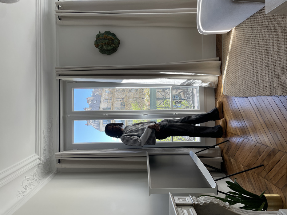 A single individual stands inside a well-lit room, facing left toward a tall window. The person has dark skin and long dark dreadlocks or braids, wearing a white long-sleeved undershirt, a light-colored plaid short-sleeved top, dark trousers, and light brown boots. Their right hand is placed in a pocket. The room features a herringbone parquet floor and a light-colored woven rug. White walls are topped with ornate decorative ceiling molding. To the left of the window, a white-framed television on a minimalist black stand is positioned next to a plant in a gold pot. To the right of the window, a green-patinated sculptural wall hanging is affixed. Sunlight streams through the window, illuminating parts of the floor and furniture. Through the window and beyond an ornate wrought-iron balcony railing, several classic Haussmannian buildings with light stone facades, multiple windows, and a distinctive mansard roof are visible across a street, characteristic architecture of Paris, France, under a clear blue sky.