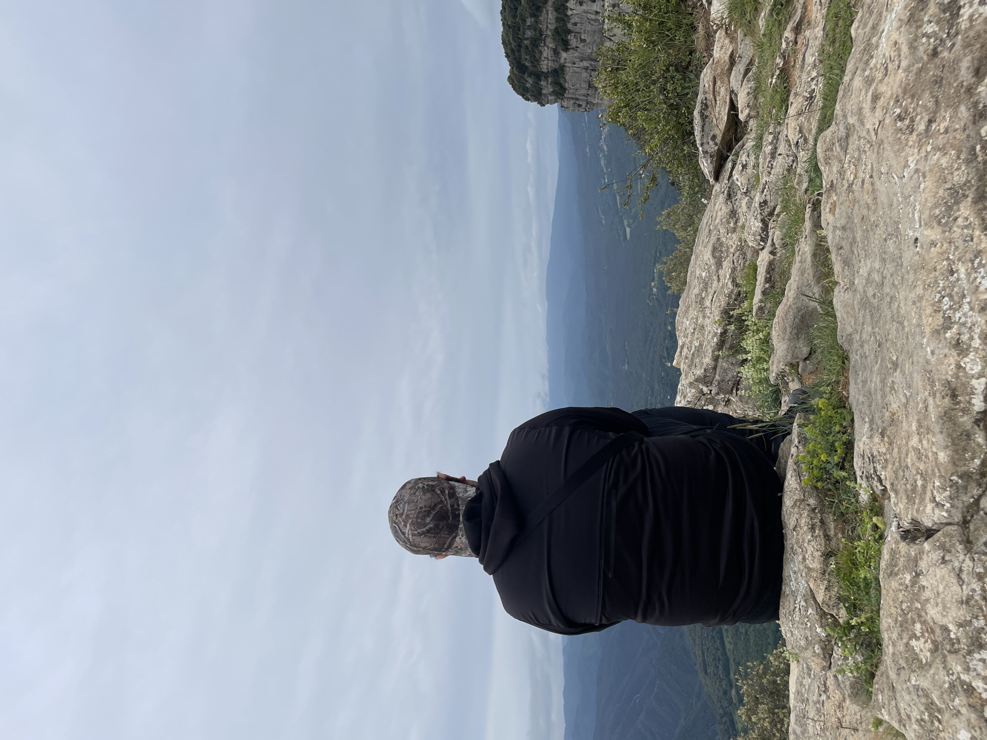 A person wearing a black hooded jacket and camouflage hat sits on the edge of a rocky cliff overlooking a mountainous landscape under a cloudy sky. The location is Tavertet, Spain, as evidenced by the terrain and vegetation. The person is facing away from the camera, looking out at the landscape. The cliff edge is characterized by exposed rock and sparse vegetation.