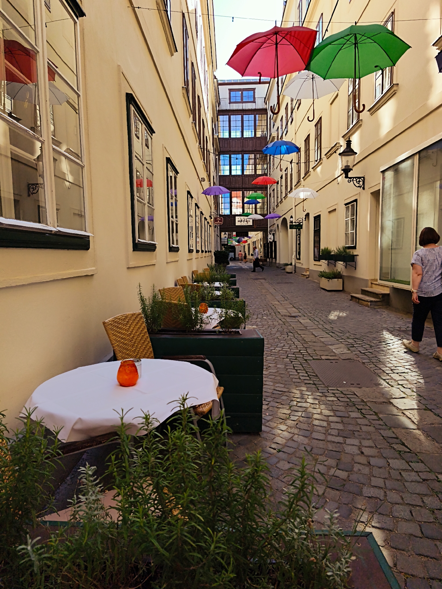 The image depicts a narrow, cobblestone street in Vienna, Austria, lined with buildings. The street is adorned with numerous colorful umbrellas suspended overhead, creating a vibrant canopy. On the left side of the street, there is an outdoor seating area with a round table covered with a white tablecloth, a woven chair, and planters filled with greenery. On the right side, a woman with dark hair is walking away from the camera, wearing a patterned blouse and dark pants. The buildings feature large windows and traditional Viennese architecture. The sunlight casts shadows on the cobblestones, suggesting it is daytime.