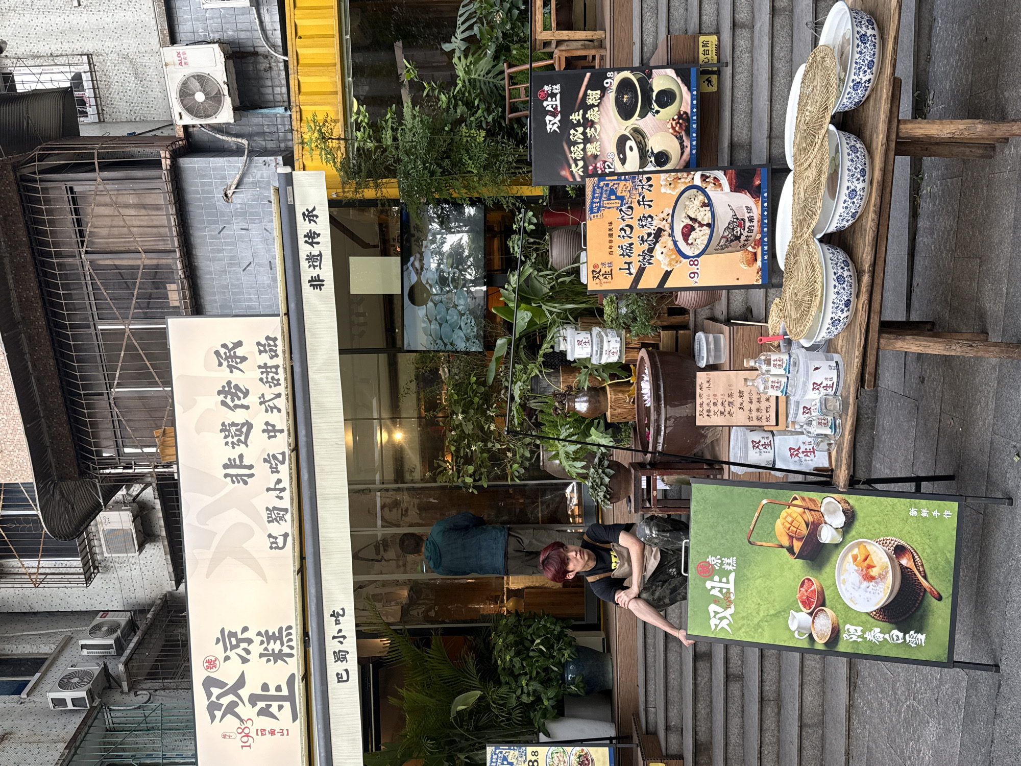 This street-level image captures the storefront of "Shuangsheng Lianggao," a dessert and snack shop, in an urban setting typical of Yuzhong District, China. The scene features a mix of traditional Chinese food culture and modern commercial display.
The main subject is a person with red hair, wearing a dark shirt and apron, leaning against a promotional A-frame sign on the sidewalk. They appear to be an employee or owner of the shop, possibly taking a break or tending to the outdoor display. Another person is partially visible inside the shop through the glass windows.
Prominently displayed are multiple signs advertising the shop's offerings. A large vertical sign on the left reads "双生凉糕" (Shuangsheng Lianggao), "非遗传承" (Intangible Cultural Heritage), "巴蜀小吃 中式甜品" (Bashu Snacks, Chinese Desserts), and "1983," suggesting its establishment year. Several A-frame signs showcase specific desserts: one features "太极双生 黑芝麻糊" (Tai Chi Shuangsheng Black Sesame Paste) for ¥12.8 and "山城记忆 双生凉糕" (Mountain City Memory Shuangsheng Lianggao) for ¥9.8. Another sign promotes "新鲜手作 阳春白雪" (Freshly Handmade Yangchun Baixue) with mango and coconut.
A rustic wooden display table holds three large, decorative blue and white ceramic bowls, possibly containing ingredients like grains or rice, topped with woven mats. Bottles of water or other beverages are also arranged on the table. Potted plants add greenery around the entrance. The building itself has large glass windows on the lower level, reflecting the surroundings, and an older, tiled facade on the upper level with air conditioning units, one bearing the "AUX" brand. The ground is paved, with steps leading up to the shop. The overall activity centers around the shop promoting and selling its traditional Chinese desserts and snacks.