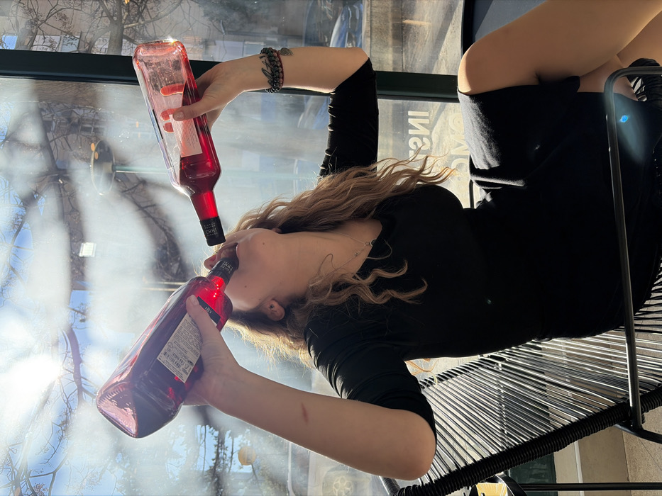 A female individual with long, wavy blonde/light brown hair is depicted seated in a black woven chair, simultaneously drinking from two clear glass bottles filled with a red liquid. Her head is tilted back, with both bottles inverted towards her open mouth, each held by one hand. She wears a black dress with three-quarter sleeves, a silver necklace with a small pendant, and a multi-beaded bracelet on her right wrist. Two linear scars are visible on her left forearm.
The scene is indoors, brightly lit by sunlight entering through a large glass window or wall. The background seen through the window includes bare tree branches, a blue sky, and distant structures, with a partial view of a white vehicle to the left. The words "INSTAGRAM MONTENEGRO" are clearly visible, mirrored in reverse, on the window's surface.
The location is identified as **Instagram Cafe, Budva, Montenegro**, based on the prominent signage reflected on the window. The environment suggests a modern cafe or bar setting with large windows.