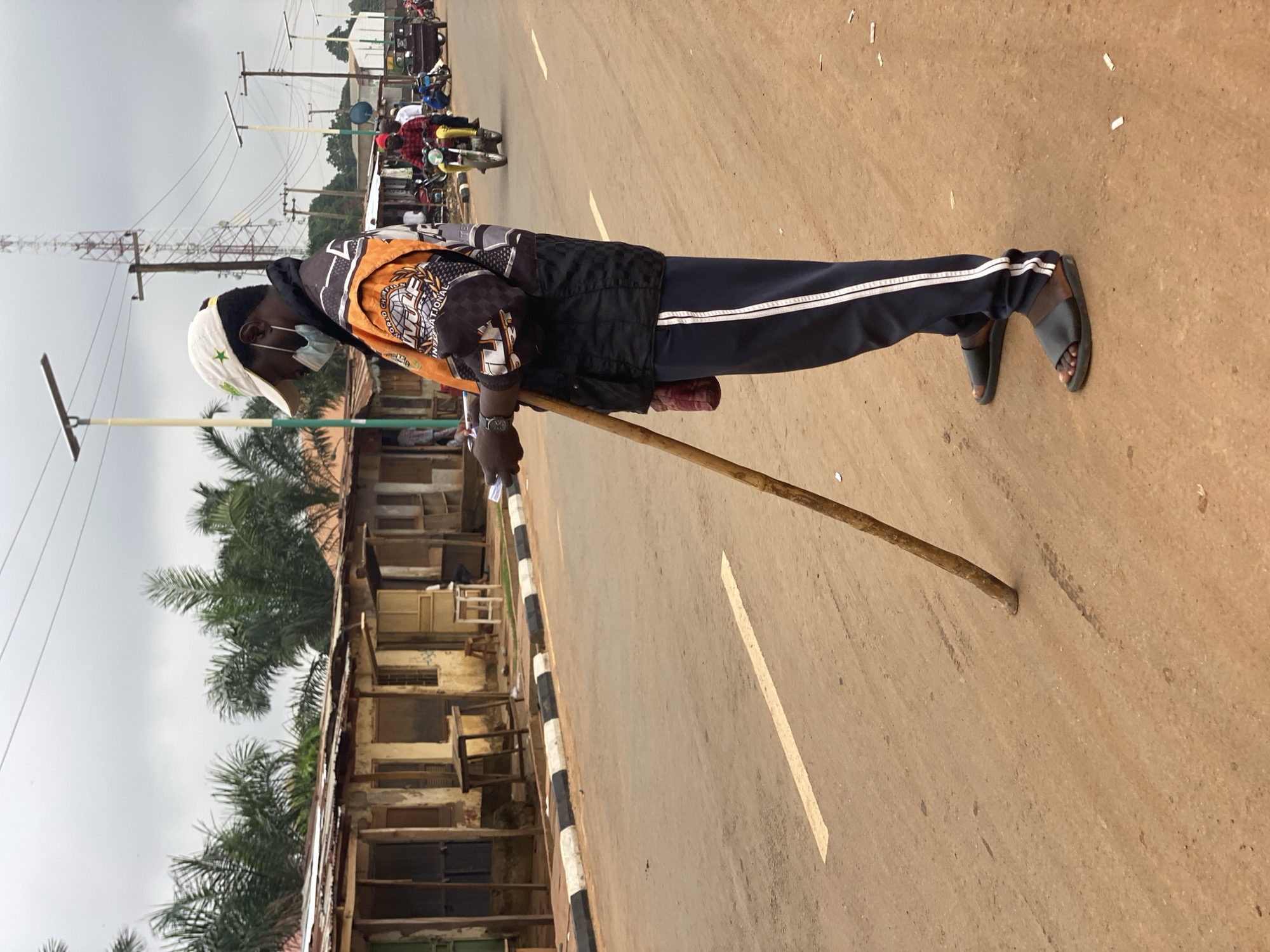 A man stands on the side of a road in Kafanchan B, Nigeria, using a wooden stick to maintain balance. He wears a white cap with a green star, a black vest over an orange and black jacket, navy blue pants with white stripes, and black sandals. He also wears a light blue face mask. Behind him, a multi-story structure is visible, with stacked wooden frames resembling windows or shelves. In the distance, utility poles, wires, and a tall metal tower are seen against a cloudy sky. Motorcycles and other figures are blurred further down the road. The road has a yellow center line and a striped black and white curb.