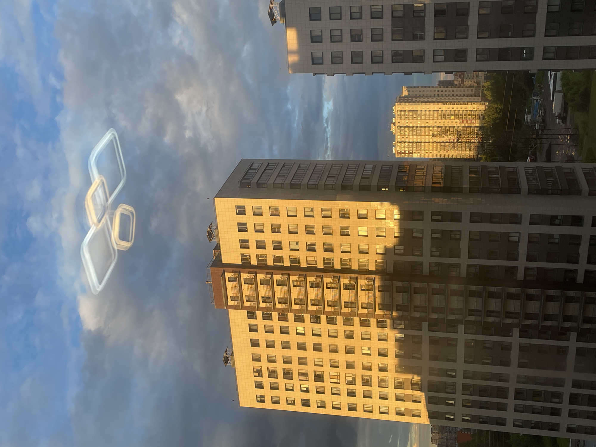 This media file captures an urban scene dominated by tall residential buildings bathed in the warm glow of the setting sun. The perspective is from a window or balcony, looking upwards and outwards.
The primary subjects are several large, modern apartment buildings. Their facades are adorned with numerous windows, reflecting the golden light. One building in the foreground is particularly detailed, showing balconies and the geometric pattern of its construction. In the background, another building, possibly older or of a different architectural style, is also illuminated by the sun. The sky above is a dramatic mix of blue and dark, cloudy formations, suggesting dynamic weather.
Suspended in the sky are three abstract, glowing, ring-like objects, possibly a form of artistic or digital overlay. They appear translucent and are clustered together, creating a surreal element against the realistic urban backdrop.
The time of day is clearly late afternoon or early evening, indicated by the low angle of the sun and the long shadows and warm lighting it casts on the buildings. The weather appears to be transitioning, with both clear sky and overcast clouds visible.
The scene evokes a sense of urban living and the beauty of natural light interacting with architecture. There is no visible text in the image. The location is not explicitly identified, but the context suggests an urban environment in Russia.