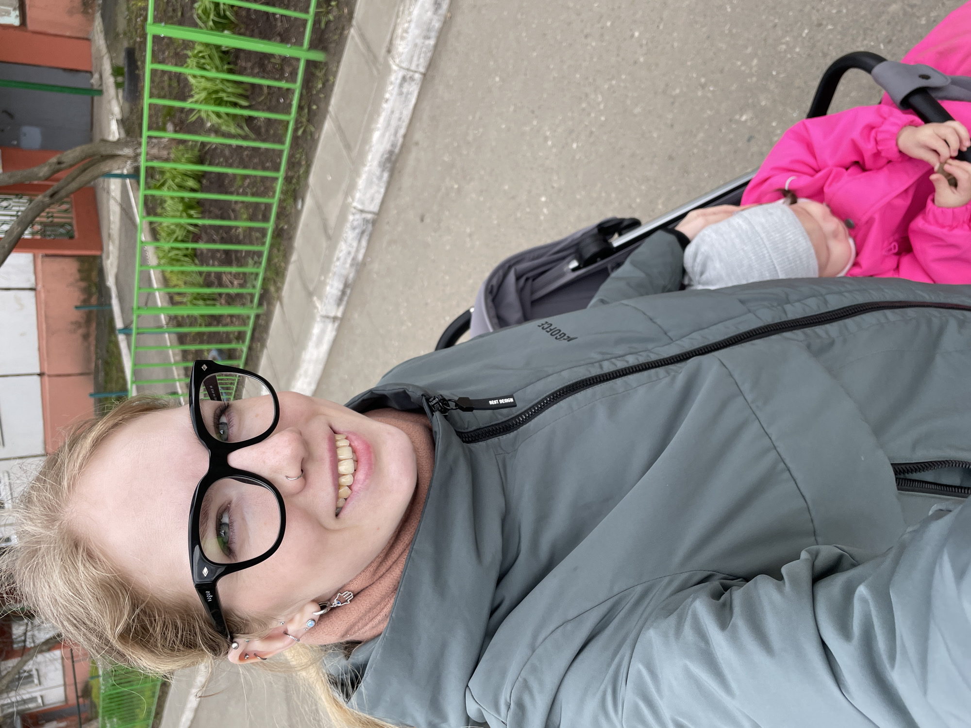 A selfie taken outdoors depicts a woman with blonde hair, fair skin, and glasses, smiling towards the camera. She wears a gray jacket over a peach-colored turtleneck. In the background, two children are visible in a stroller. One child is wearing a pink jacket and the other a gray hat. The scene appears to be on a sidewalk next to a building, which has a green fence. Based on the architecture and green metal fence, this appears to be in Izhevsk, Russia.
