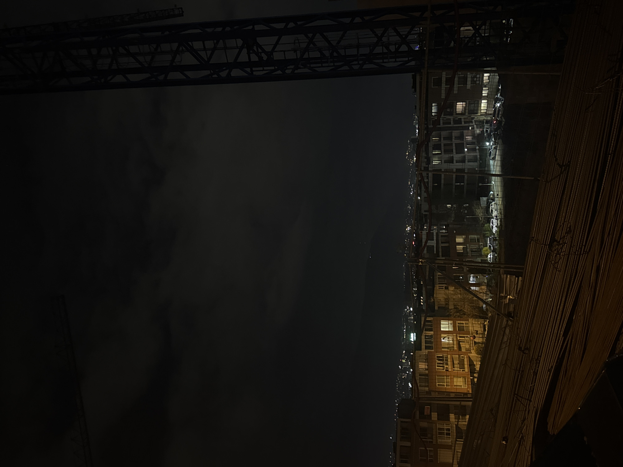 A low-angle nighttime shot shows a city landscape in Sofia, Bulgaria, partially obscured by a large metal structure in the upper left and what appears to be stacked construction materials in the lower right. The city buildings, lined with lit windows, stretch into the distance, illuminated against the dark sky. Car headlights indicate street activity.
