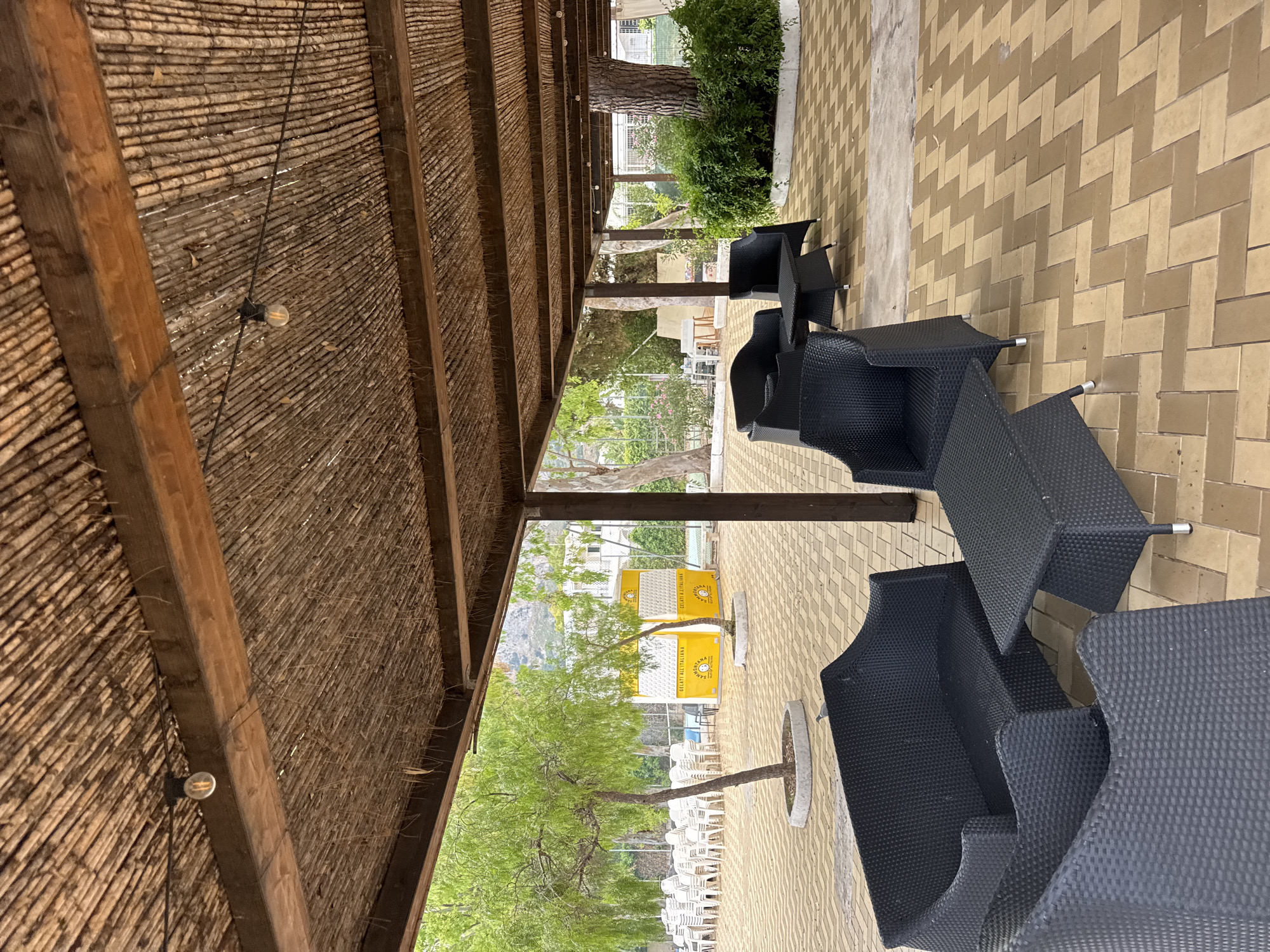 The image shows an outdoor seating area under a thatched roof pergola. The pergola has wooden beams and is illuminated by string lights with exposed bulbs. In the foreground, there are several pieces of black wicker furniture, including a sofa and armchairs, arranged around black wicker coffee tables. The floor is paved with light brown and tan hexagonal tiles laid in a herringbone pattern.
In the middle ground, there are two yellow structures with "GELATI ALL'ITALIANA" branding, suggesting they are ice cream stands. A small tree is planted between them. In the background, rows of white plastic chairs are visible, indicating a potential event or dining area. Beyond that, there are buildings and lush green trees. A mountain range is visible in the distance. The overall setting appears to be a public space in Isola delle Femmine, Italy, possibly a beach resort or park.