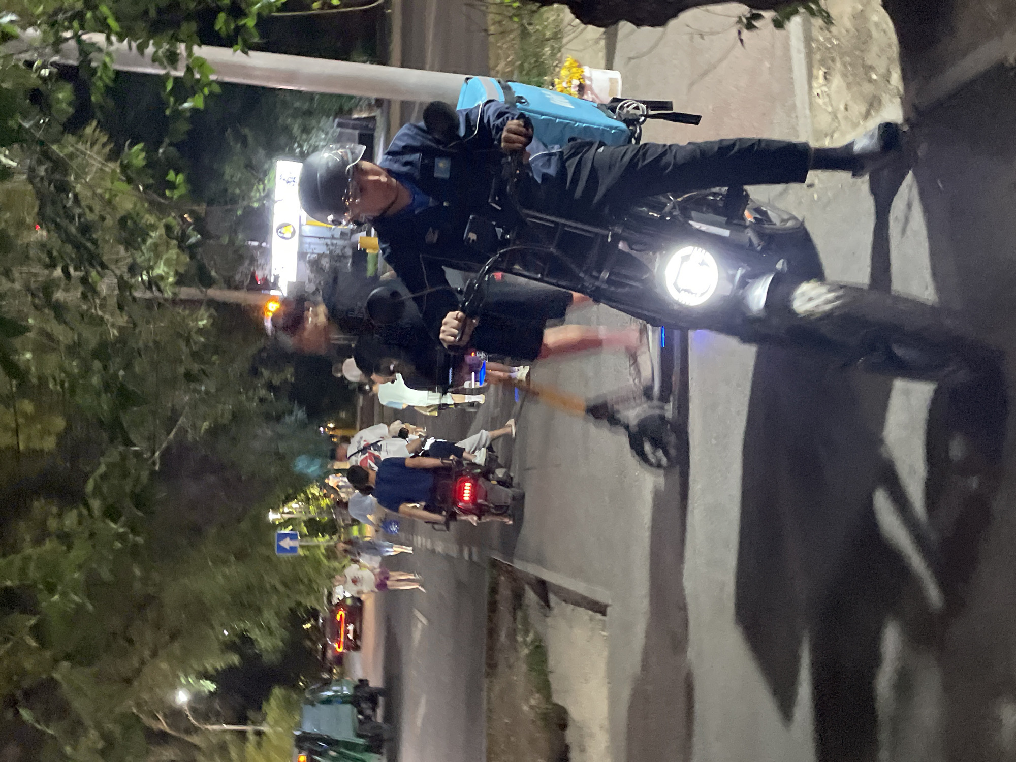 In Almaty, Kazakhstan, at night, a person on a motorcycle is the primary focus. The rider, wearing a dark blue jacket with a Kazakh flag patch on the sleeve and a helmet with a clear visor, is positioned in the foreground, facing slightly to the left. The motorcycle has a bright headlight on and a large, light blue insulated delivery bag is attached to the rear. Behind the motorcycle, a sidewalk is visible with several other people walking and standing. One person is on a scooter, and another is riding a red motorcycle with illuminated taillights. In the background, trees line the street, and distant streetlights and building lights illuminate the urban environment. A red car is visible on the road to the left. The scene appears to be a street or pathway in a residential or commercial area.