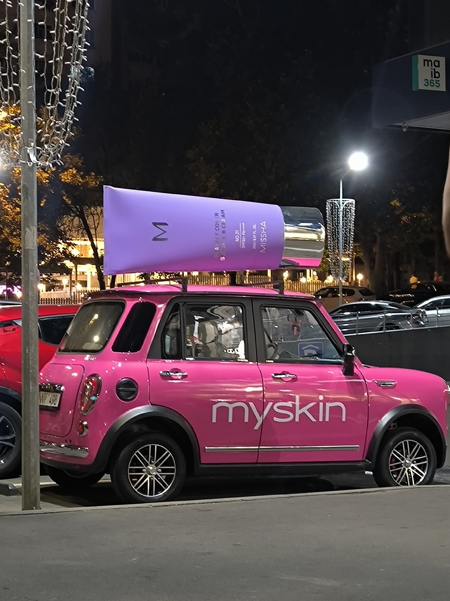 A bright pink car with the brand name "myskin" emblazoned on its side is parked at street level in an urban environment at night. The car is a modern, compact vehicle with chrome accents and alloy wheels. Perched on the roof rack of the car is a large, oversized replica of a purple cosmetic tube, featuring the "M" logo and text that appears to be "MISSHA." The car is situated next to other vehicles, including a red car parked to its left, and appears to be on a paved surface, possibly a road or parking lot. In the background, there are trees, buildings, and streetlights, suggesting a city setting. A sign with "ma ib 365" is visible in the upper right corner, indicating a commercial establishment nearby. The overall scene conveys a promotional event or advertising display for a cosmetic product. The location is identified as Chișinău, Moldova.