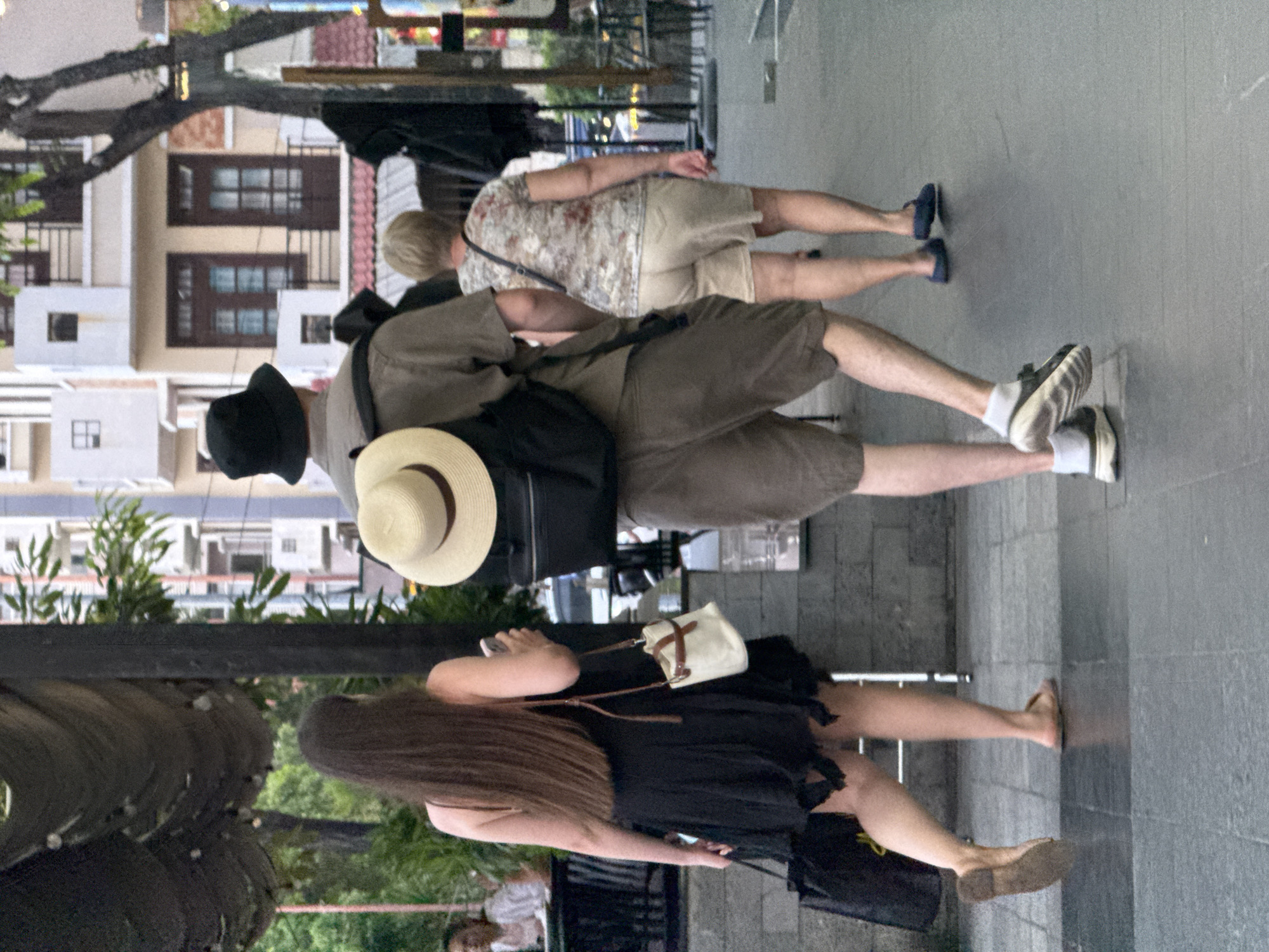 The image captures a street scene in Nha Trang Ward, Vietnam, featuring three individuals walking on a paved sidewalk. In the foreground, a woman with long, dark hair, wearing a black sleeveless dress and a small cream-colored crossbody bag, walks from left to right. She is wearing a flat brown sandal on her left foot and her right foot is partially visible. She is carrying a black tote bag in her left hand. To her right, a man wearing a light brown t-shirt, a bucket hat, and dark brown shorts walks towards the viewer, carrying a black backpack. He is wearing white socks and dark athletic shoes. Behind him, further to the right, a woman with short blonde hair, wearing a patterned t-shirt and light-colored shorts, also walks towards the viewer. She has a dark bag slung over her shoulder. The background shows buildings with balconies and windows, some with awnings, and trees. The lighting suggests it is daytime.