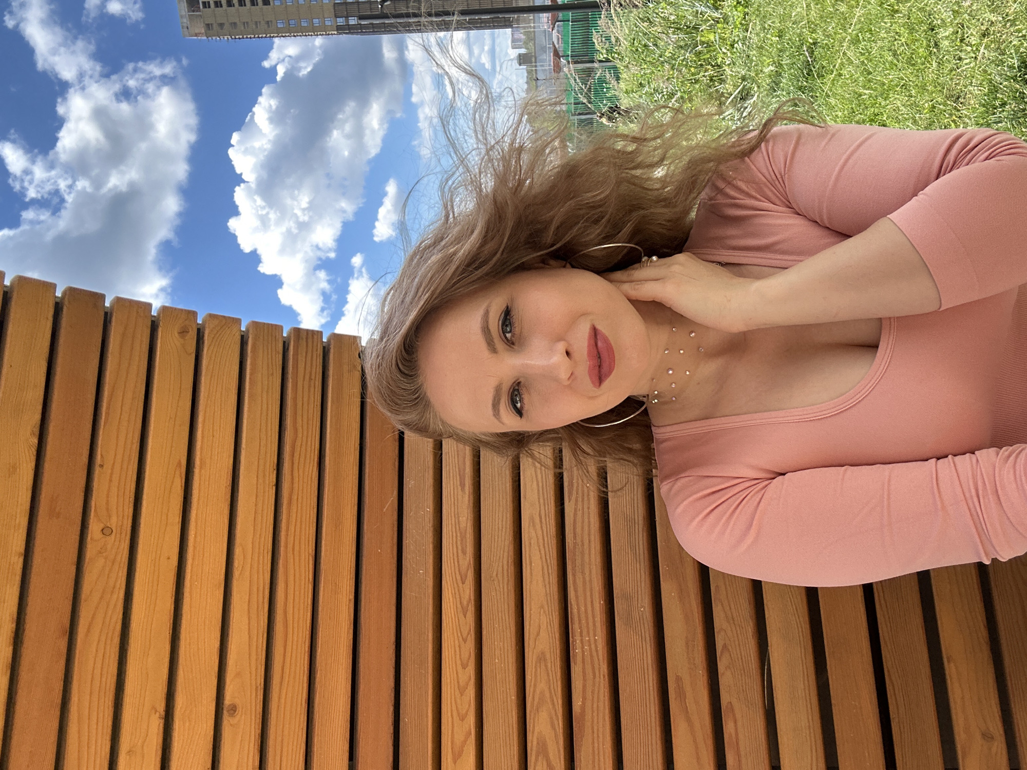 A medium shot shows a woman with wavy blonde hair and fair skin, wearing a pink long-sleeved shirt, and a necklace of small rhinestones. She is positioned in front of a wooden slatted wall. Her right hand is touching her neck. The background features a green grassy area, a fence, a cloudy blue sky, and a tall, unfinished building. The scene is located in the city of Krasnogorsk, Russia.