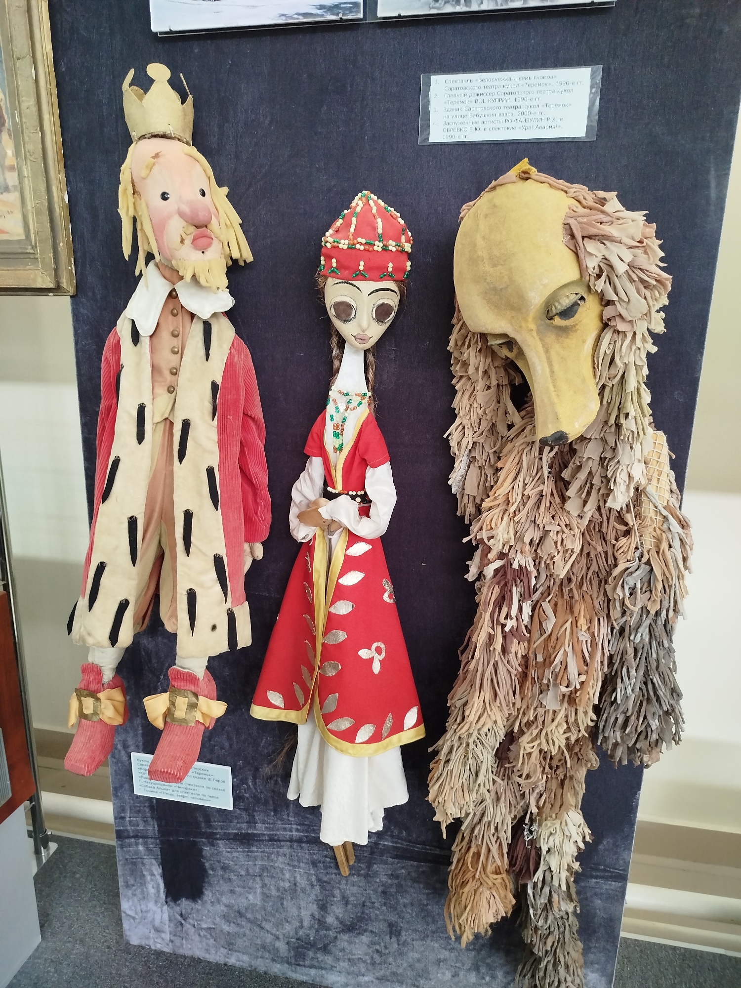 Three puppets are displayed against a dark blue backdrop. On the left, a male puppet resembling a king wears a red robe lined with fur trim and a crown. He has a pale, elongated face with large eyes, a prominent nose, and a beard. To the right of the king puppet is a female puppet dressed in a red and white costume adorned with beads and embroidery. She has a slender neck and large, dark eyes. On the far right, a puppet with a yellow, mask-like face is covered in shaggy, textured material that resembles fur or long hair. A small white sign with Russian text is attached to the backdrop. The puppets appear to be part of an exhibit, possibly in a museum or theater.