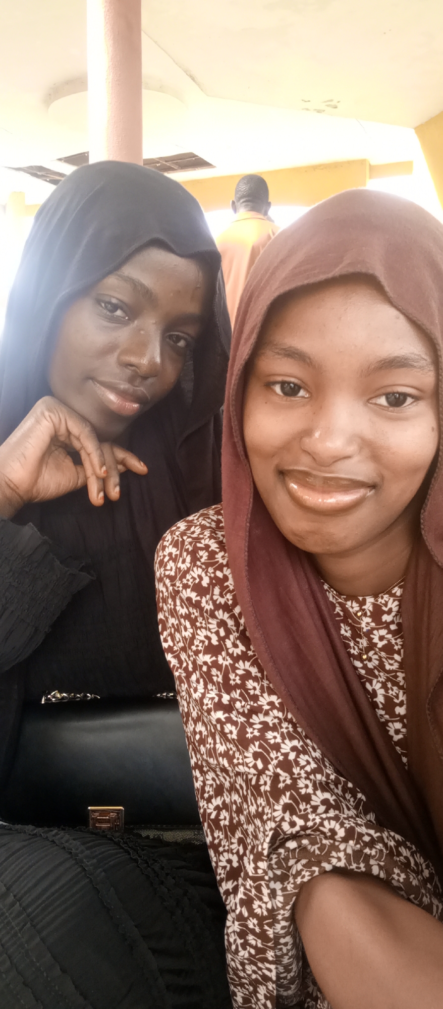 Two young women are prominently featured in a photo, likely a selfie, smiling directly at the camera. The setting appears to be indoors or under a covered outdoor structure, characterized by a bright, light-colored ceiling and a visible pillar in the background, suggesting daytime.
The woman on the left has dark skin and wears a black hijab and a black dress with pleated or ruffled sleeves. She has a gentle smile, and her right hand, with fingers stained reddish-brown (possibly with henna), rests on her chin. A black handbag is visible in the lower left portion of the image. The woman on the right, also with dark skin, wears a brown hijab and a brown dress adorned with a white floral pattern. She sports a wide, cheerful smile.
In the blurred background, a man wearing an orange or yellow shirt is partially visible from behind, facing away from the camera. There is no visible text in the image. The overall mood is pleasant and friendly. This scene is located in Keffi, Nigeria.