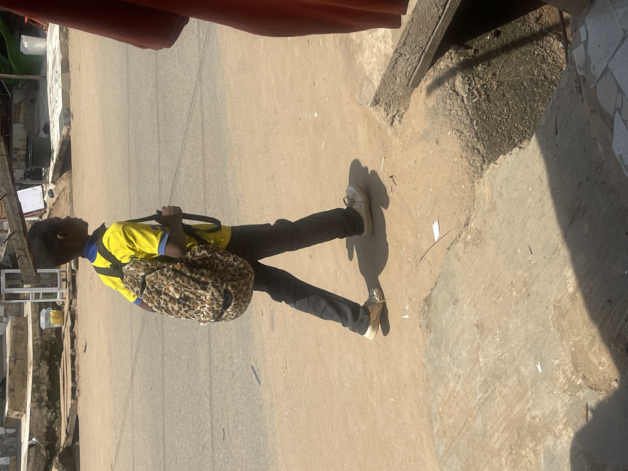 A young boy walks on a dirt road in Abuja, Nigeria, carrying a leopard-print backpack. He is wearing a yellow polo shirt with blue trim and dark pants. The boy is walking from left to right in the image, and his shadow is visible on the ground. The background shows a street with some buildings and debris. The sun is shining brightly, casting strong shadows.
