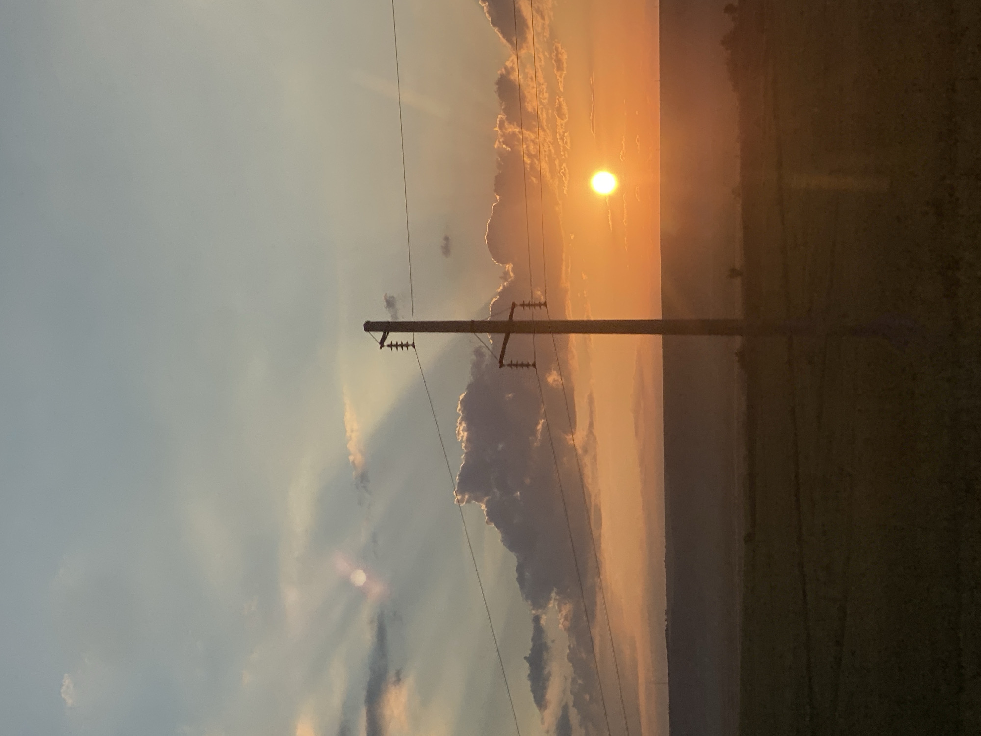 The image depicts a sunset scene in Balabanu, Moldova. A single utility pole stands in the foreground, supporting electrical wires that stretch across the frame. The sun, a bright orange orb, is setting on the horizon, casting a warm glow across the sky and illuminating scattered clouds. A vast, dark field stretches out below the horizon, suggesting agricultural land. The overall impression is one of rural tranquility at dusk.