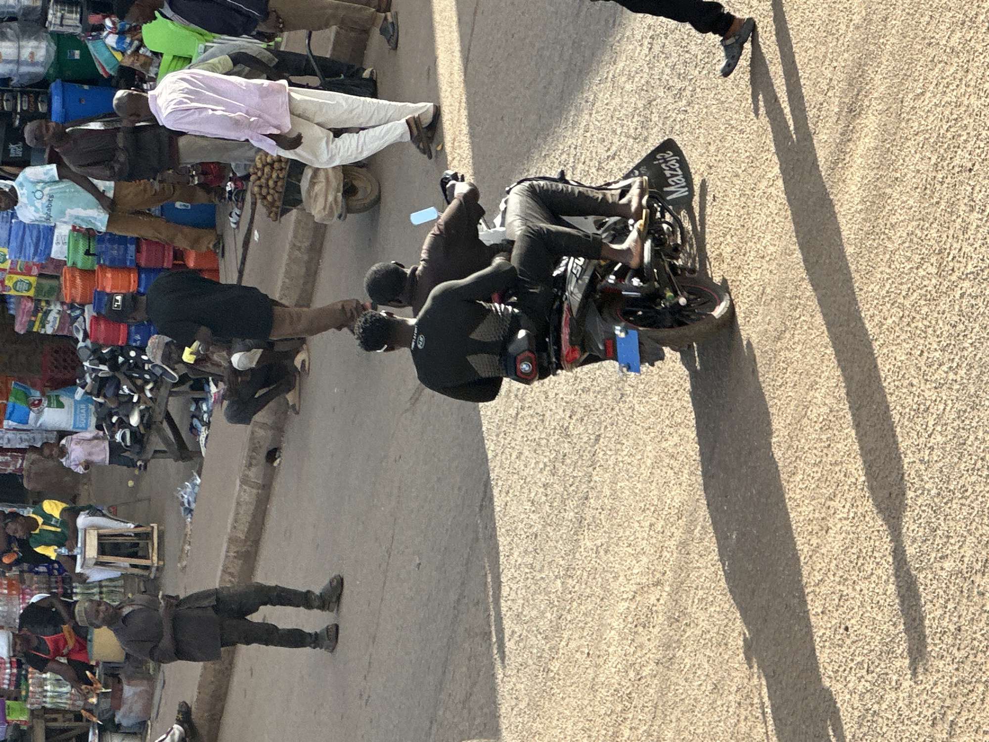 The image depicts a bustling street scene in Bukuru, Nigeria. Several people are visible, some walking, others on a motorcycle. The primary subjects are individuals engaged in daily activities within a market or commercial area. One person is wearing a light pink shirt and white pants, standing near a stall. Another group is gathered around a motorcycle, with one person appearing to be the rider. The motorcycle itself is black and red, with the word "Mazajaz" visible on its rear fender in white lettering.
The setting appears to be a street lined with various goods, suggesting a marketplace. Crates and merchandise are visible on the left side of the image, including what looks like bottles and bags of produce. The ground is paved with asphalt and gravel. The lighting indicates it is daytime, with strong shadows cast on the ground, suggesting a time around midday or early afternoon. The weather appears to be sunny.
The overall atmosphere seems lively and active, typical of a busy commercial hub. The people's postures and proximity suggest movement and interaction. No specific emotions are overtly displayed, but the scene conveys a sense of ordinary commerce and transit. The presence of a motorcycle with passengers and the surrounding market stalls points to a functional urban environment where people are going about their business.