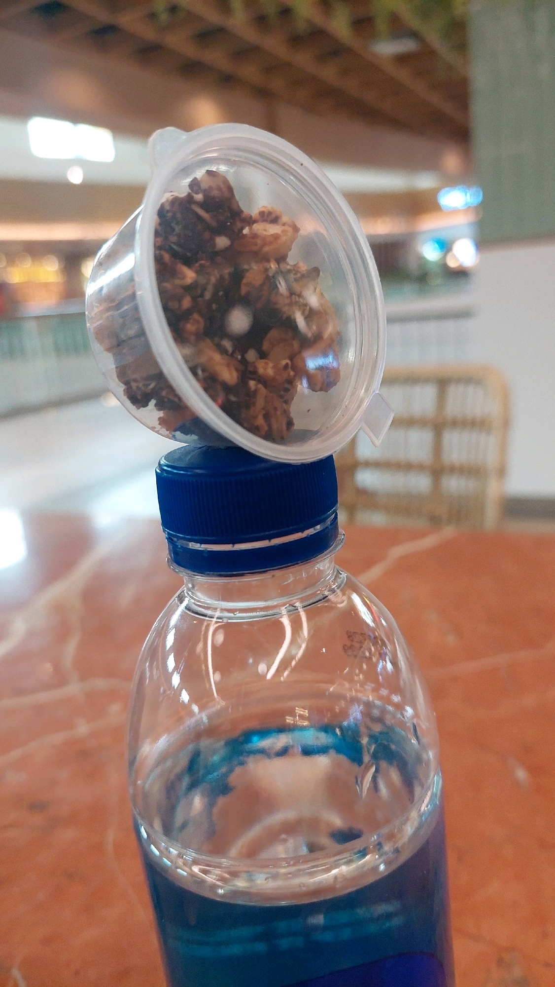 A clear plastic bottle with blue liquid is on a reddish-brown surface. A small, clear plastic container filled with dark granola-like material is balanced on the blue cap of the bottle. The background is blurred, showing what appears to be an indoor environment with wooden ceiling beams and out-of-focus lights and furniture, possibly in a shopping mall in North Jakarta, Indonesia.
