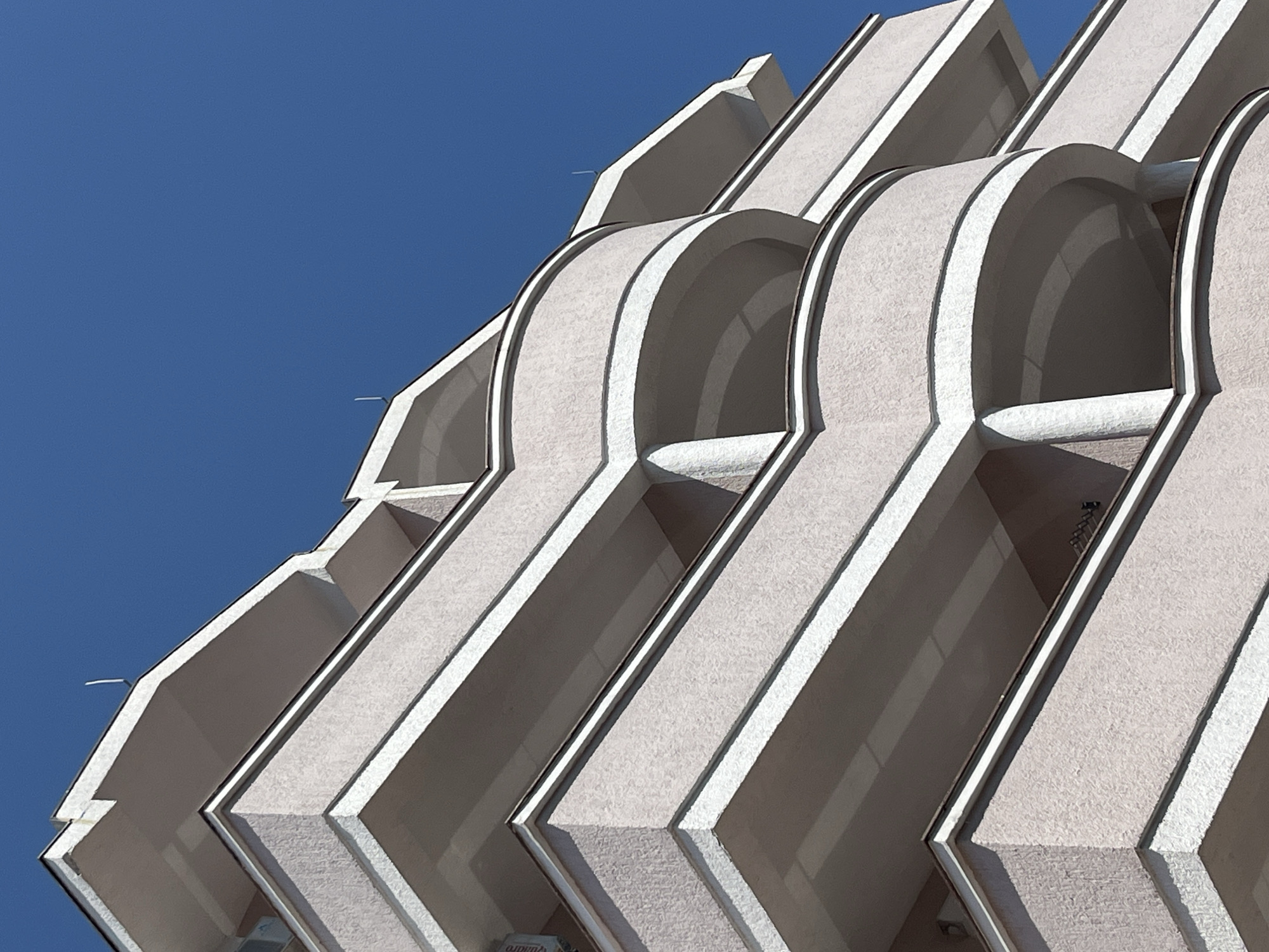 This is a low-angle, close-up, abstract shot of the exterior of a modern building, likely an apartment complex or hotel, against a clear blue sky. The building's facade features a repeating pattern of recessed, arched openings, framed by sharp, angular white trim. The material of the facade appears to be textured and light-colored, possibly concrete or stucco. The repeating architectural elements create a sense of rhythm and depth in the composition. Sunlight casts strong shadows, highlighting the three-dimensional nature of the design and creating a dynamic interplay of light and dark. The clear sky suggests a daytime setting, likely with good weather. No people or specific objects are clearly visible. The image focuses on the architectural details of the building itself. Based on the provided location context, this building is located in Budva, Montenegro.