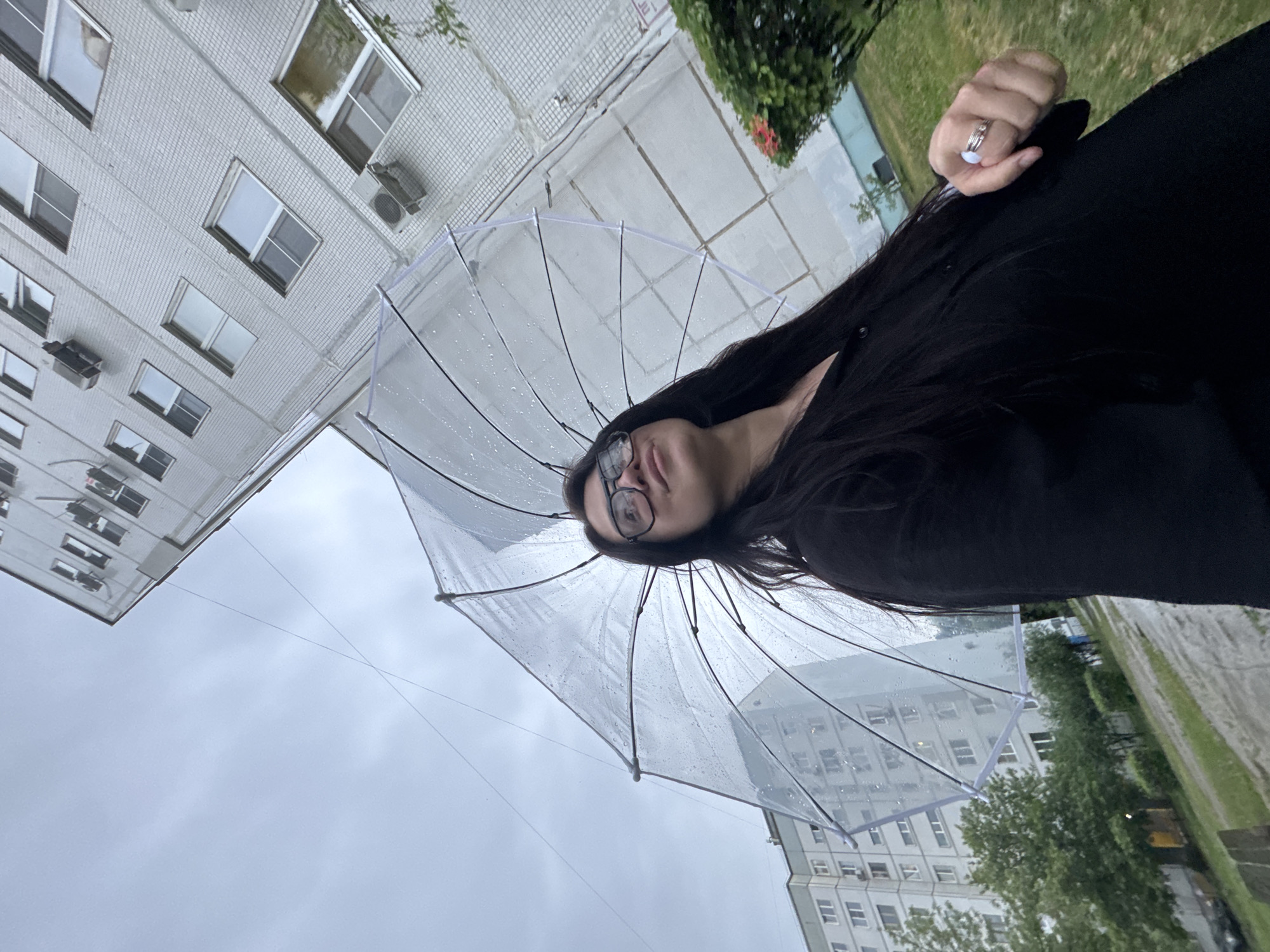 A low-angle, rotated-perspective shot reveals a woman holding a clear umbrella, with buildings and a cloudy sky visible through it. The woman has long, dark hair, wears glasses, and is dressed in dark clothing. She is holding the umbrella with her right hand, and rings are visible on her fingers. The umbrella is wet with raindrops. Reflected and obscured through the clear canopy of the umbrella is an indistinct building. Outside the boundaries of the umbrella frame, there is also a view of trees and grass. The sky is overcast.