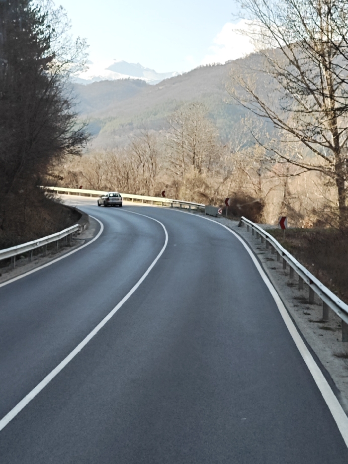 The image depicts a two-lane asphalt road featuring an S-curve, situated in a mountainous environment identified as Gospodintsi, Bulgaria. The dark grey road surface is marked by a continuous white line separating the two lanes and solid white lines along the outer edges. Galvanized metal guardrails are present along both sides of the road's curves. Multiple red and white chevron signs are visible, indicating the presence of sharp turns. A single light-colored hatchback vehicle is observed traveling away from the viewer, positioned in the right lane within the second segment of the S-curve. The surrounding terrain is covered with deciduous trees, predominantly bare of leaves, suggesting a late autumn or winter season. Distant mountain peaks, some snow-capped, are visible under a light blue sky with minimal cloud cover.