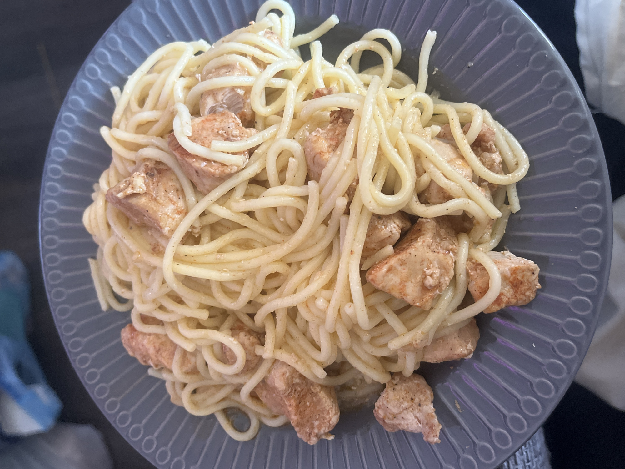 A grey, circular ceramic plate, featuring a raised, repetitive linear and circular pattern along its rim, contains a serving of light-colored spaghetti mixed with cuboid pieces of cooked chicken. The chicken exhibits a reddish-brown exterior with specks of dark seasoning, suggesting it has been pan-fried or baked. The spaghetti strands are coated in a pale, creamy-looking sauce, with some darker seasoning visible on them as well. The plate rests on an indistinct, dark surface. The background is out of focus, revealing a blurry light blue object to the upper left and general darker tones. The location of this scene in the city of Unknown, Russia, cannot be determined from the visual information provided.