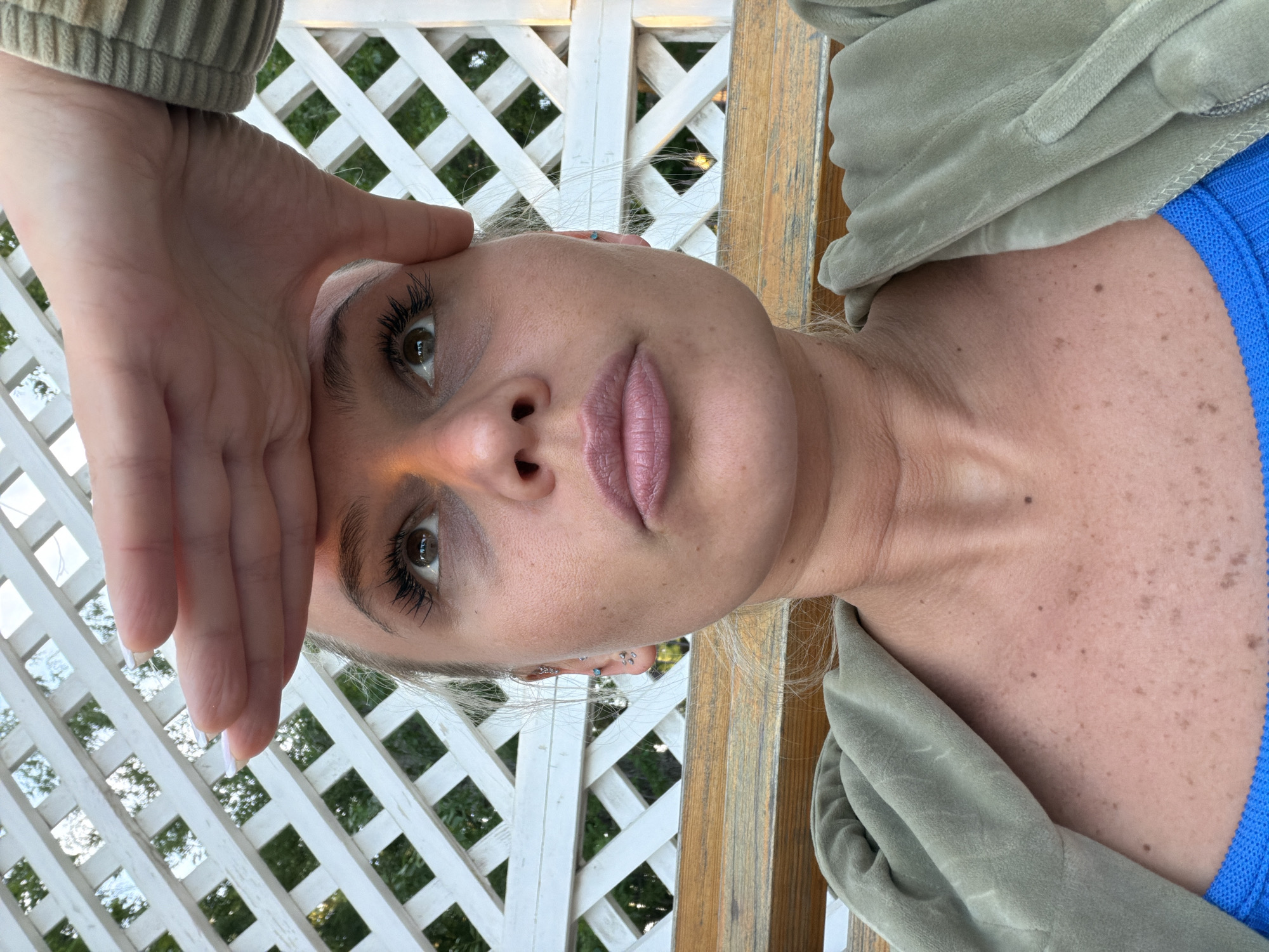 A light-skinned female with blonde hair poses close to the camera. Her hand rests on her forehead. She has brown eyes and pink lips. The background features a white lattice fence and vegetation. The environment provides no clear indication of the city of Sarapul, Russia. She is wearing a blue shirt and a light green jacket. The photograph appears to be taken outdoors in natural light.